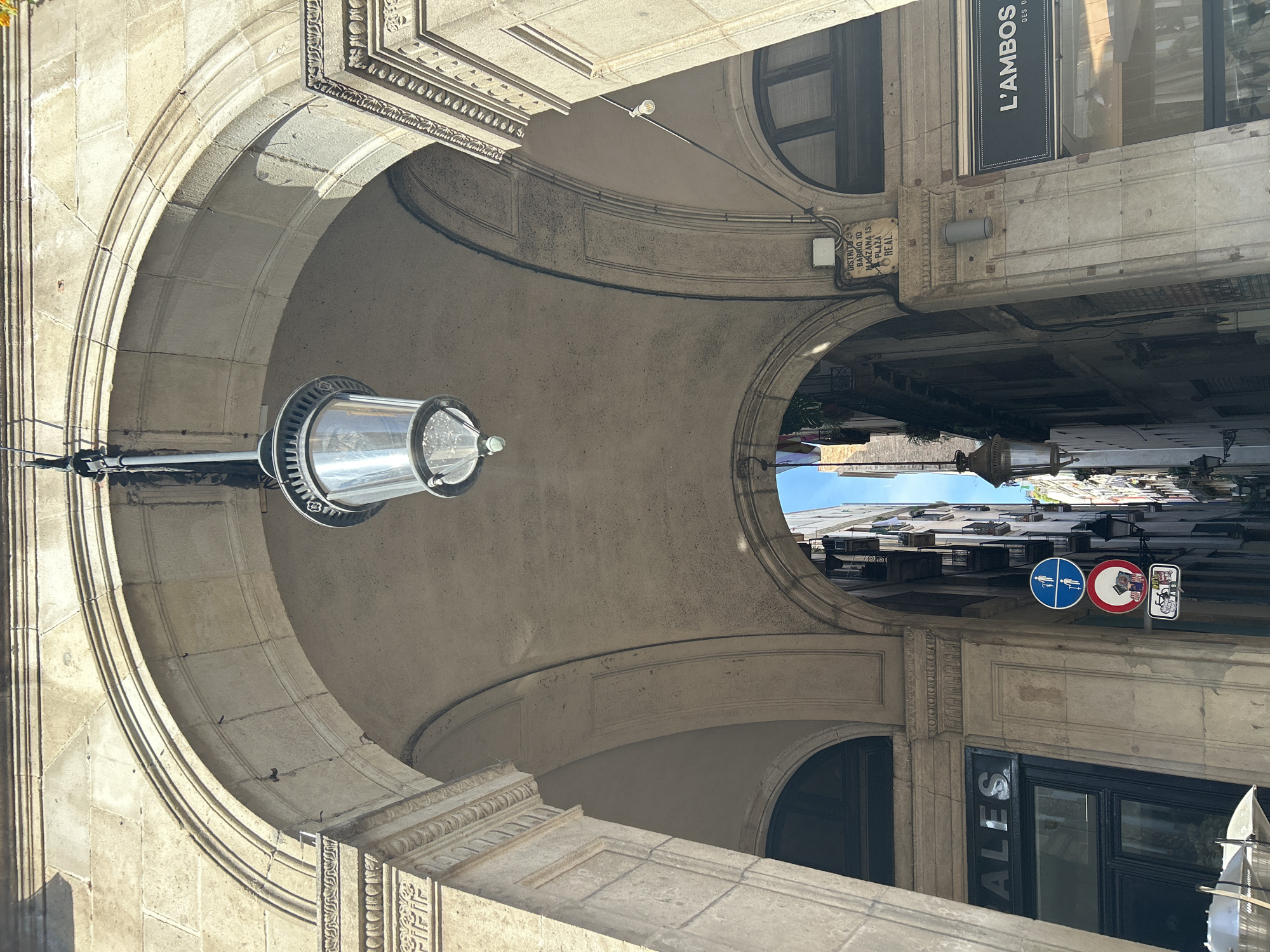 The image shows an archway in Barcelona, Spain, with a street lamp hanging on the left. The archway is made of stone with decorative moldings. A sign on the right reads "L'AMBOS." The street sign indicates a walking path and prohibition. The view through the archway reveals buildings and a sliver of blue sky. There is a plaque that is in district 10 of Barcelona in the Marzana district on Plaza Real.