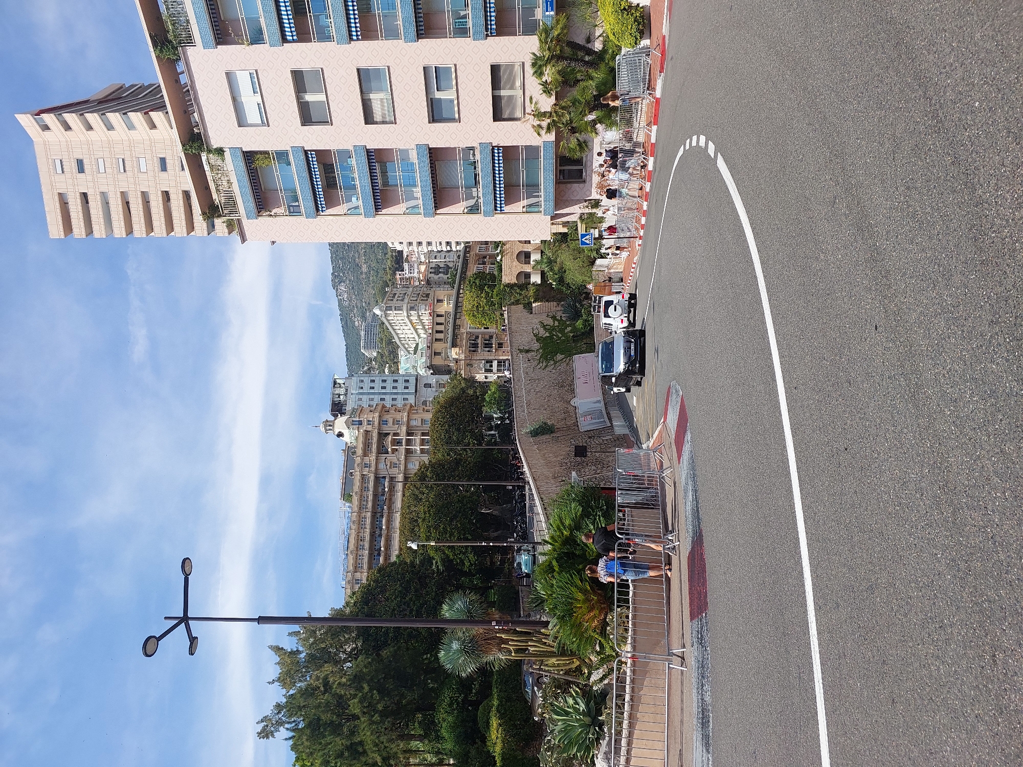 The image depicts a street scene in Monaco, likely on a race circuit given the barriers and the road markings. In the foreground, a paved road curves to the right, marked with a white line. A metal barrier with a red and white striped base runs along the edge of the road, separating it from a landscaped area. Several people are visible standing behind the barrier, observing the scene.
Behind the barrier, a lush garden features various cacti, palm trees, and other greenery. A tall streetlamp with three lights stands on the left side of the image. Further in the background, several buildings rise up the hillside. The most prominent is a tall, modern, light-colored skyscraper. To its left, a pink building with blue awnings and balconies is visible. Beyond these, numerous other buildings of varying heights and architectural styles are clustered on the slopes, suggesting a densely populated urban environment. The sky is blue with some scattered white clouds. Two cars are on the road: a silver sedan turning into the curve and a white SUV further up the road.