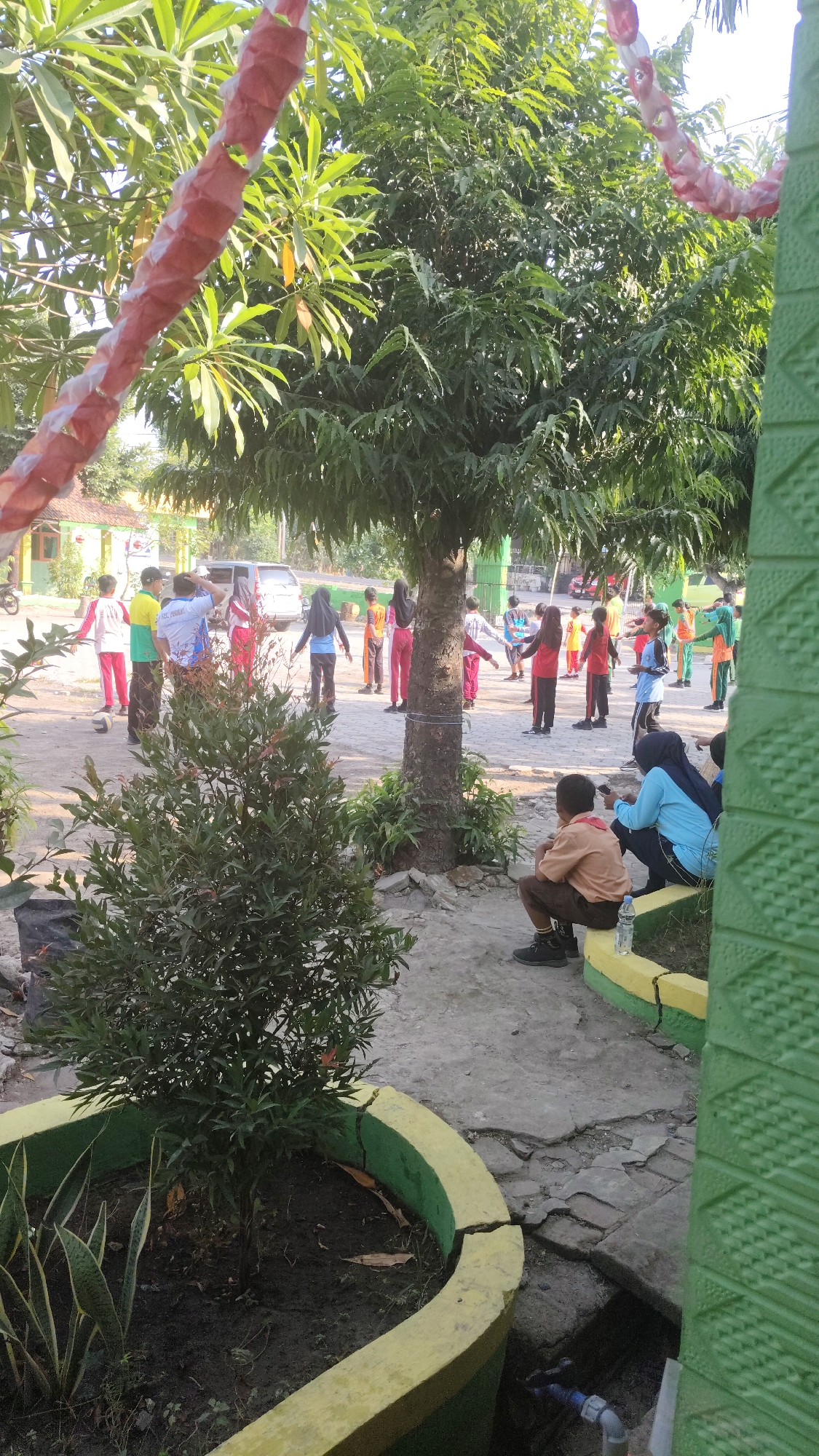 In Banjarsari, Indonesia, a group of elementary school children are engaged in a physical education class outdoors. The children, dressed in various school uniforms including tan, blue, red, and yellow tops with matching pants, are spread across a paved area beneath the shade of trees. Some children are actively participating in exercises, while others, including a boy in a tan uniform sitting on the ground and a girl in a blue hijab and shirt, are observing or resting. A few adults, one wearing a black hijab, are also present, possibly supervising the activity. The environment features a cracked concrete pathway, a planter with a small tree and snake plant, and glimpses of a building with a red roof in the background. The scene is illuminated by natural daylight, suggesting it is daytime.