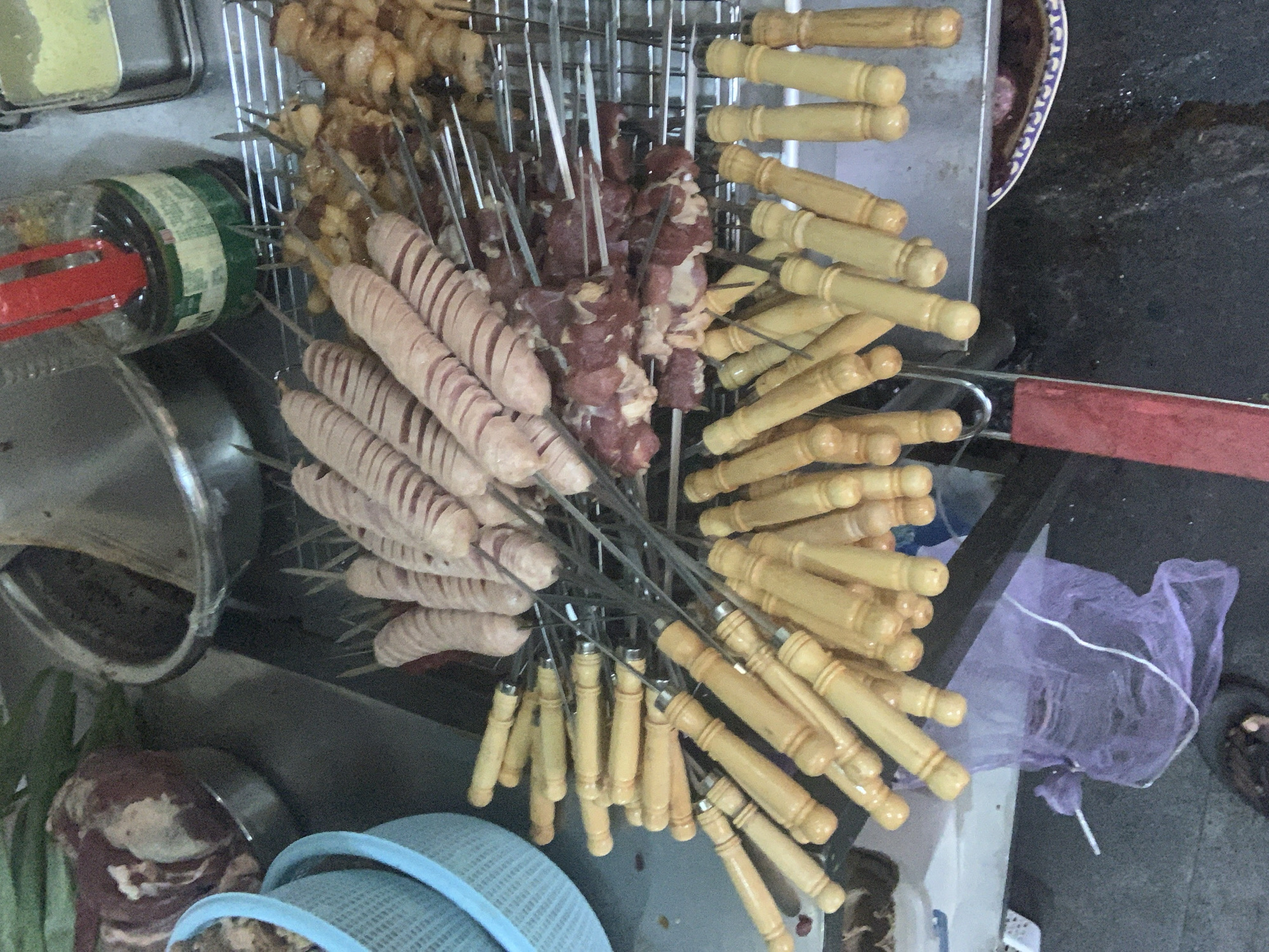 The image shows a close-up, high-angle view of skewers of raw meat and sausages arranged on a metal rack, likely at a street food stall. The skewers have wooden handles and metal prongs. In the background, a large metal bowl contains more meat, and a glass bottle with a red cap sits on a shelf. A blue plastic basket is visible on the left. The ground appears to be a dark, possibly wet, surface. The scene depicts food preparation and is not specifically identifiable as being in Abuja, Nigeria, or any other specific city.