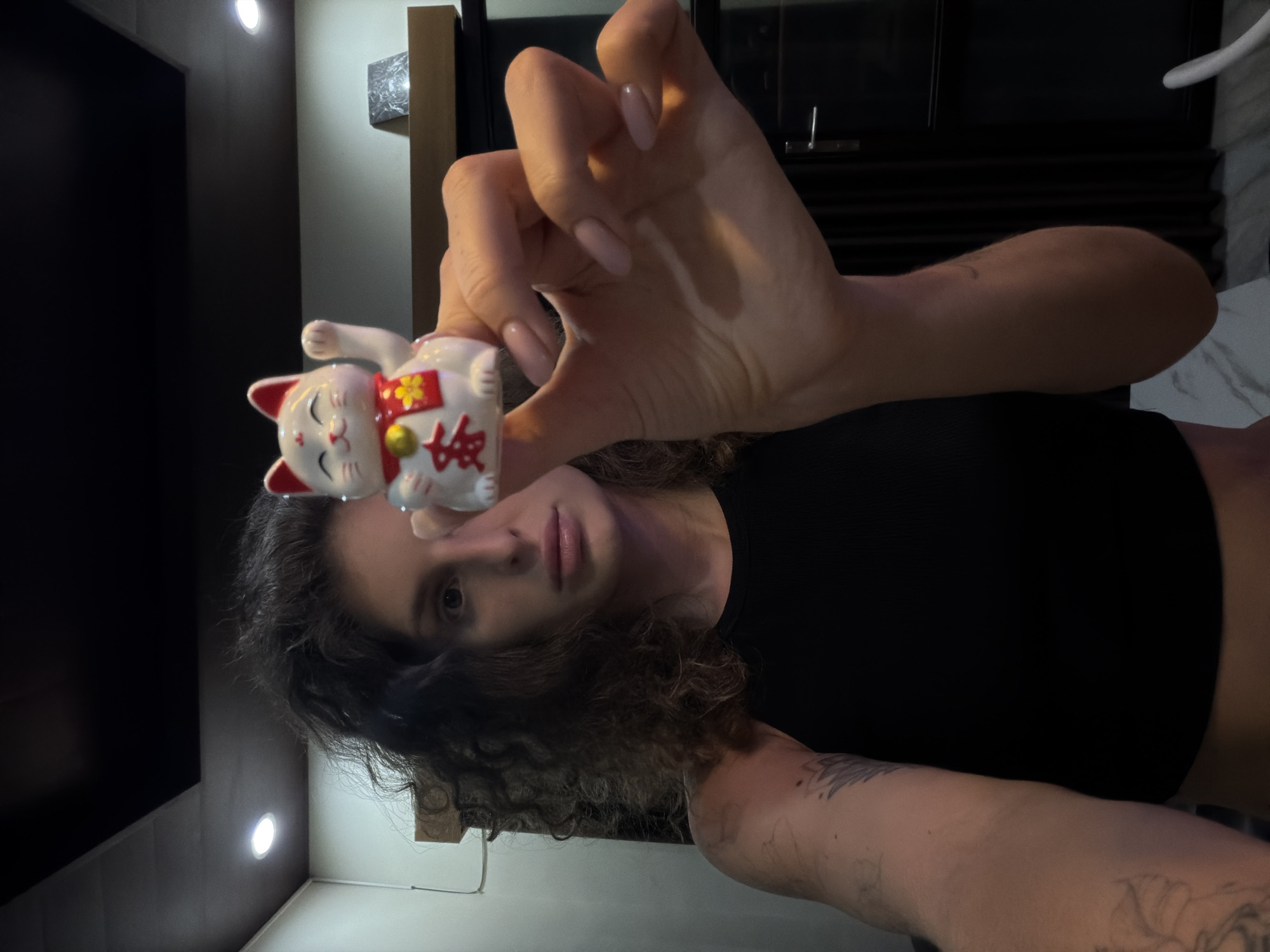 A person is depicted indoors, holding a white Maneki-neko figurine in their right hand, which is positioned in front of their face. The image is captured from a low angle, with the person looking directly at the camera. They have dark, curly hair and are wearing a black crop top. Their nails are painted a light, neutral color. Visible tattoos are on their left arm.
The Maneki-neko figurine is white with red ears, paws, and a collar. It features a smiling face with closed eyes and is adorned with a red bib that has a yellow four-petal flower design. A Japanese character, clearly "招" (sho), which is part of "招き猫" (Maneki-neko, meaning 'beckoning cat'), is prominently visible on the bib, associated with good fortune.
The background shows an indoor setting, possibly a room or hallway, with dark walls or furnishings. Bright circular ceiling lights are visible, illuminating parts of the scene. To the right, a darker vertical surface, potentially wood, is present, with a lighter, possibly marble, surface below it. The artificial lighting suggests the photograph was taken during evening or night. While the location context is given as North Kuta, Indonesia, this is not visually discernable from the image itself.