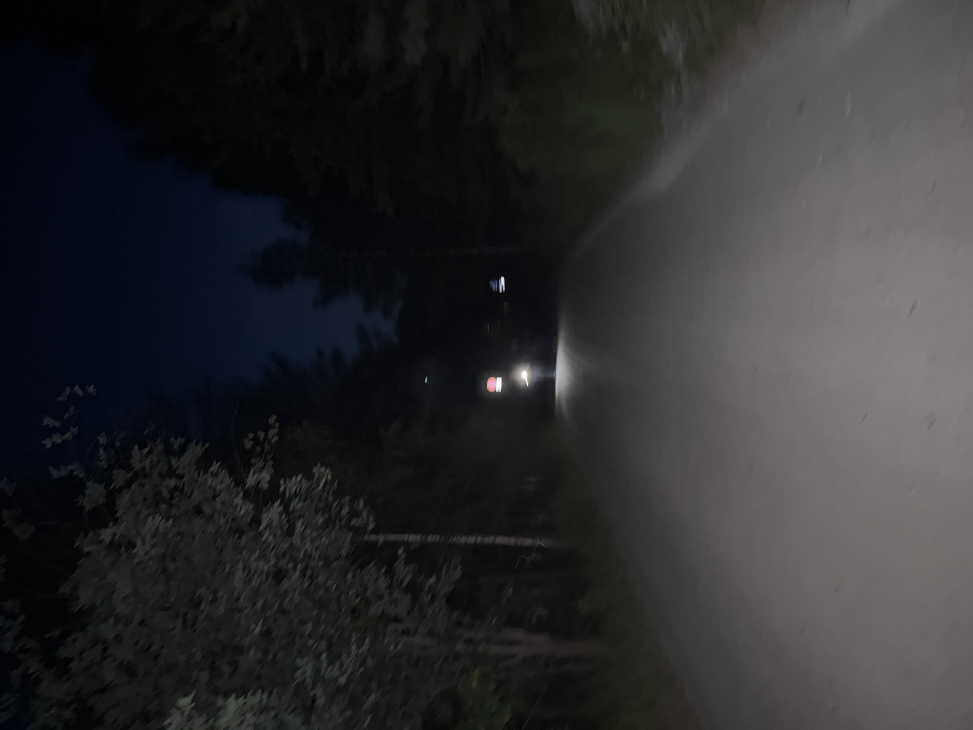 The image depicts a dirt road at night in a forested area, possibly near Dom Otdykha Zolotoy Plyazh, Russia. The road is dark and appears to be unpaved, with trees lining both sides. In the distance, a vehicle's headlights are visible, illuminating the road ahead. There are also some lights and structures on the right side of the road in the distance, which are unclear due to the low light conditions. The sky is dark, indicating it is nighttime.