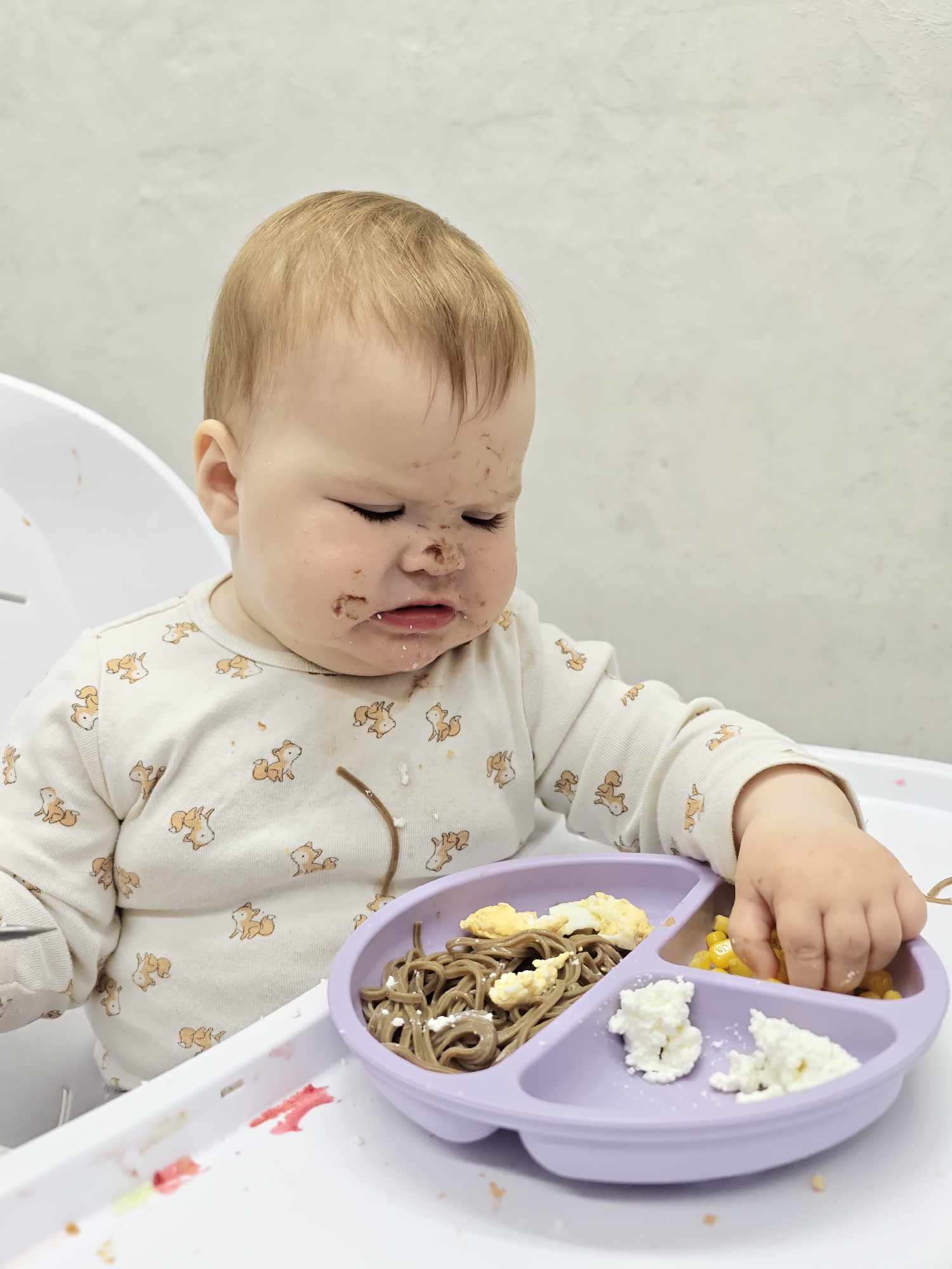 A close-up shot of a baby sitting in a high chair. The baby has light hair and is wearing a light-colored onesie with fox patterns. The baby's face is smudged with food. The baby is looking down at a purple divided plate containing noodles, corn, egg, and cottage cheese. The baby's hand is reaching into the corn section of the plate. The background is a plain white wall. The location is not specified as Chaikovsky, Russia, but the image details do not contradict that location.