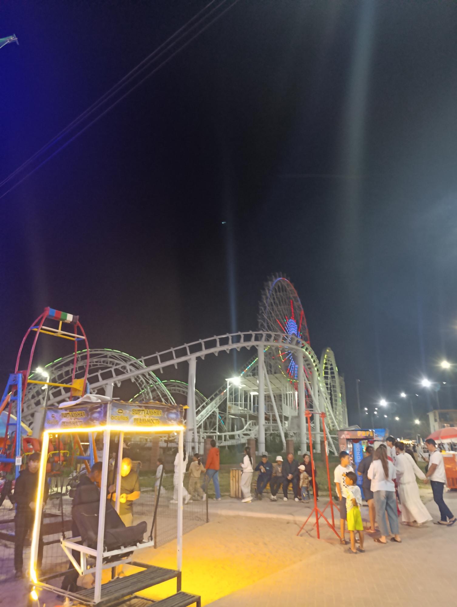 This is a night-time outdoor scene at an amusement park, likely located in Bosteri, Kyrgyzstan, given the specific context of the park. The image features a roller coaster with white tracks illuminated by green lights winding through the frame. In the background, a Ferris wheel with colorful lights (red, blue, and white) is visible, reaching high into the dark night sky.
In the foreground, a virtual reality attraction is prominent. It consists of a white metal frame supporting a seat with a black cushioned back. Bright yellow lights surround the top of this structure, casting an orange glow on the surrounding area. A few people are gathered around this attraction, some appearing to be waiting or observing.
Further to the right, a group of people are casually standing and interacting. They are dressed in a variety of casual clothing. To the left of the virtual reality setup, a smaller, colorful amusement ride with red and blue framing and a yellow bucket-like attachment is visible. The ground is paved with a textured surface, illuminated by various artificial lights from the park attractions and overhead lamps. The overall atmosphere suggests a lively evening at an amusement park.