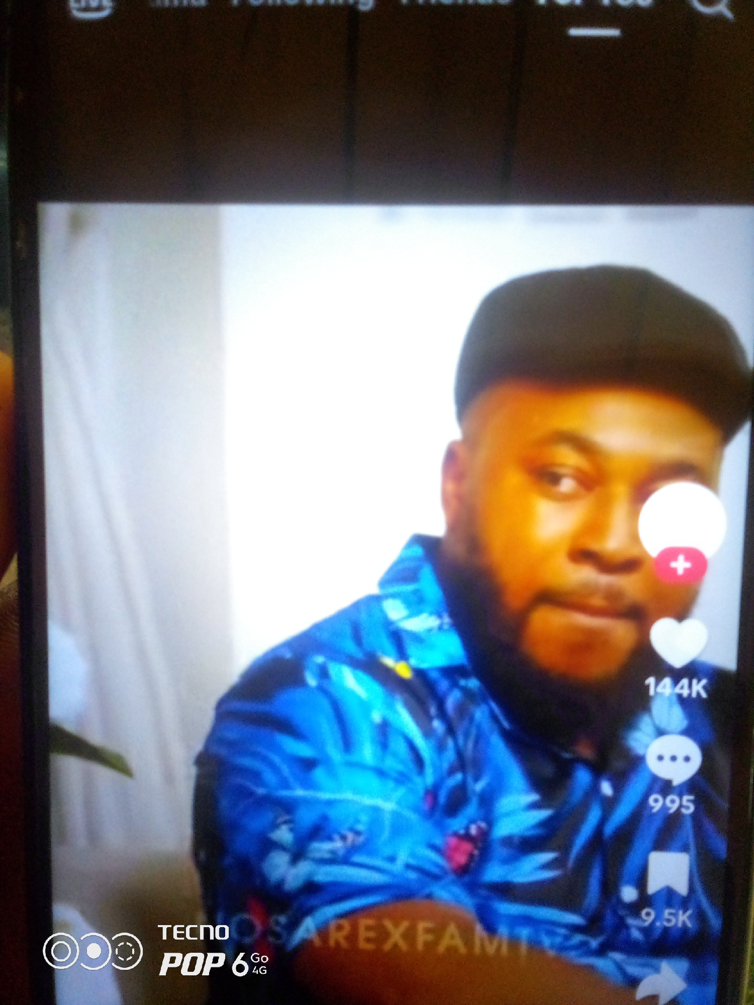 The media file shows a close-up of a Black man wearing a blue floral shirt and a black cap. He has a beard and is looking towards the camera with a neutral expression. The man is illuminated by a warm light, highlighting his face. On the right side of the image, there are social media icons and metrics: a white heart with a red plus sign, a heart icon with "144K" below it, a speech bubble icon with "995" below it, and a bookmark icon with "9.5K" below it. At the bottom left of the image, the text "TECNO POP 6 Go 4G" is visible. The scene appears to be indoors, possibly a recording studio or a home setting. There is a hint of a white background behind the subject. The overall mood is casual and relaxed. The text at the top of the image, though partially obscured, seems to be part of a social media interface, possibly indicating "Live," "Following," and "For You."