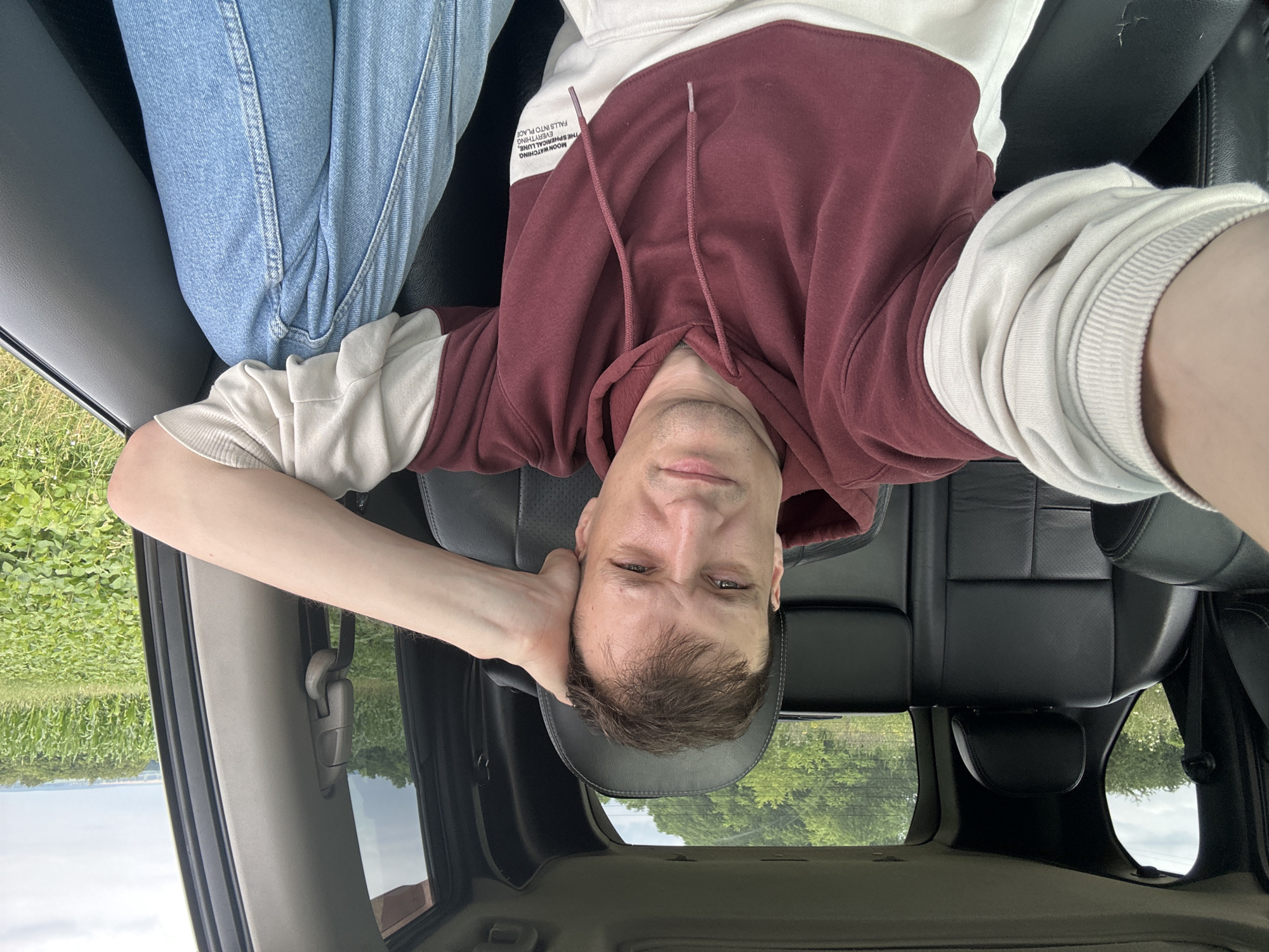 A Caucasian male, appearing to be in his late 20s or early 30s, is seated in the driver's seat of a vehicle. He has fair skin, short brown hair, and is looking towards the camera with a neutral expression. His right arm is resting on the back of his head. He is wearing a burgundy and off-white color-blocked hooded sweatshirt and light blue denim jeans. The interior of the car features black leather seats and a sunroof. Through the windows, a rural landscape is visible, with green fields, trees, and a cloudy sky. The specific location is identified as the city of Korchivka, Ukraine.