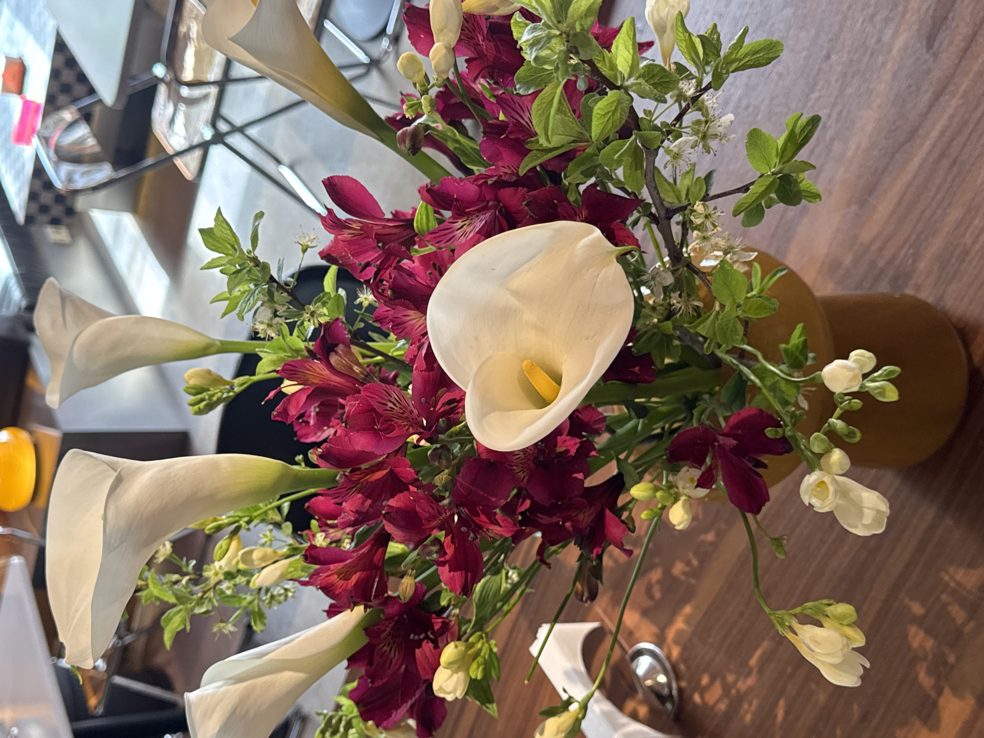 An overhead shot showcases a floral arrangement in Yerevan, Armenia, situated on a wooden table, presumably inside a building. The arrangement features white calla lilies, dark red alstroemeria, sprigs with small green leaves, and delicate white flowers. A light brown vase holds the arrangement. A glimpse of a dining area with chairs, a table, and geometric design elements on a wall is visible in the background, indicating a restaurant or cafe setting. A spoon and white napkin sit on the table near the floral arrangement.