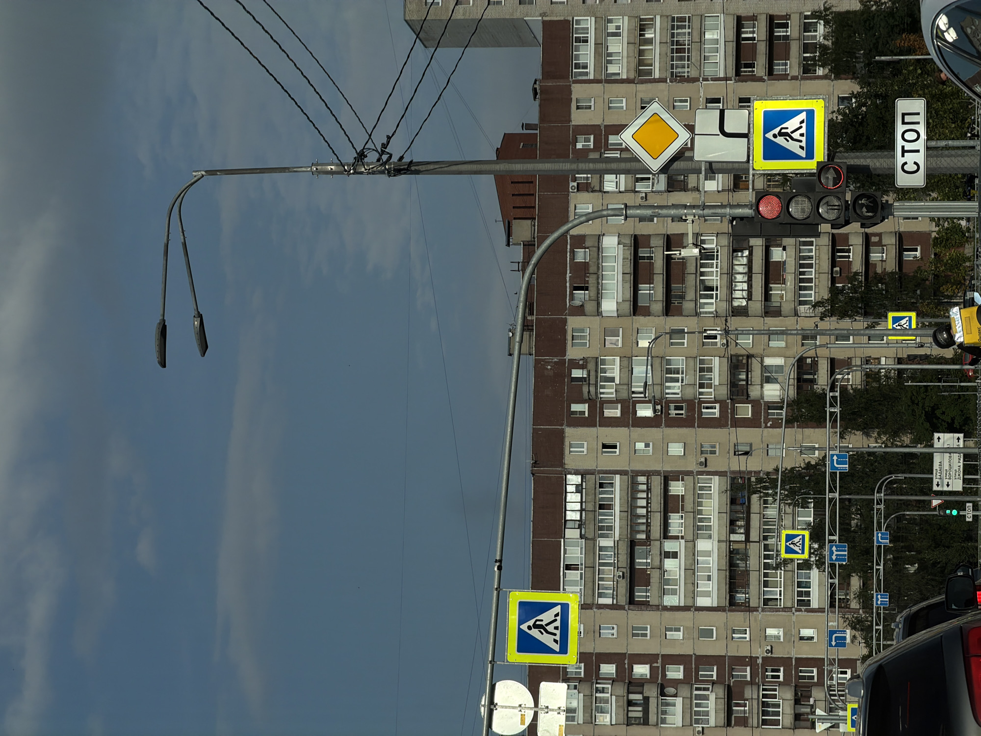 The image depicts a street scene in Saint Petersburg, Russia, dominated by a large, multi-story apartment building with a reddish-brown and beige facade. Several pedestrian crossing signs, identifiable by their blue square background with a white pedestrian silhouette, are visible. A traffic light, currently displaying red for all signals, is mounted on a pole alongside directional signs and a "STOP" sign. Power lines crisscross the upper portion of the image, and a street lamp with two lights is prominently featured. The sky is partly cloudy with a mix of blue sky and grey clouds. In the foreground, the rear of a dark car is visible. The overall impression is that of a typical urban environment in Russia.