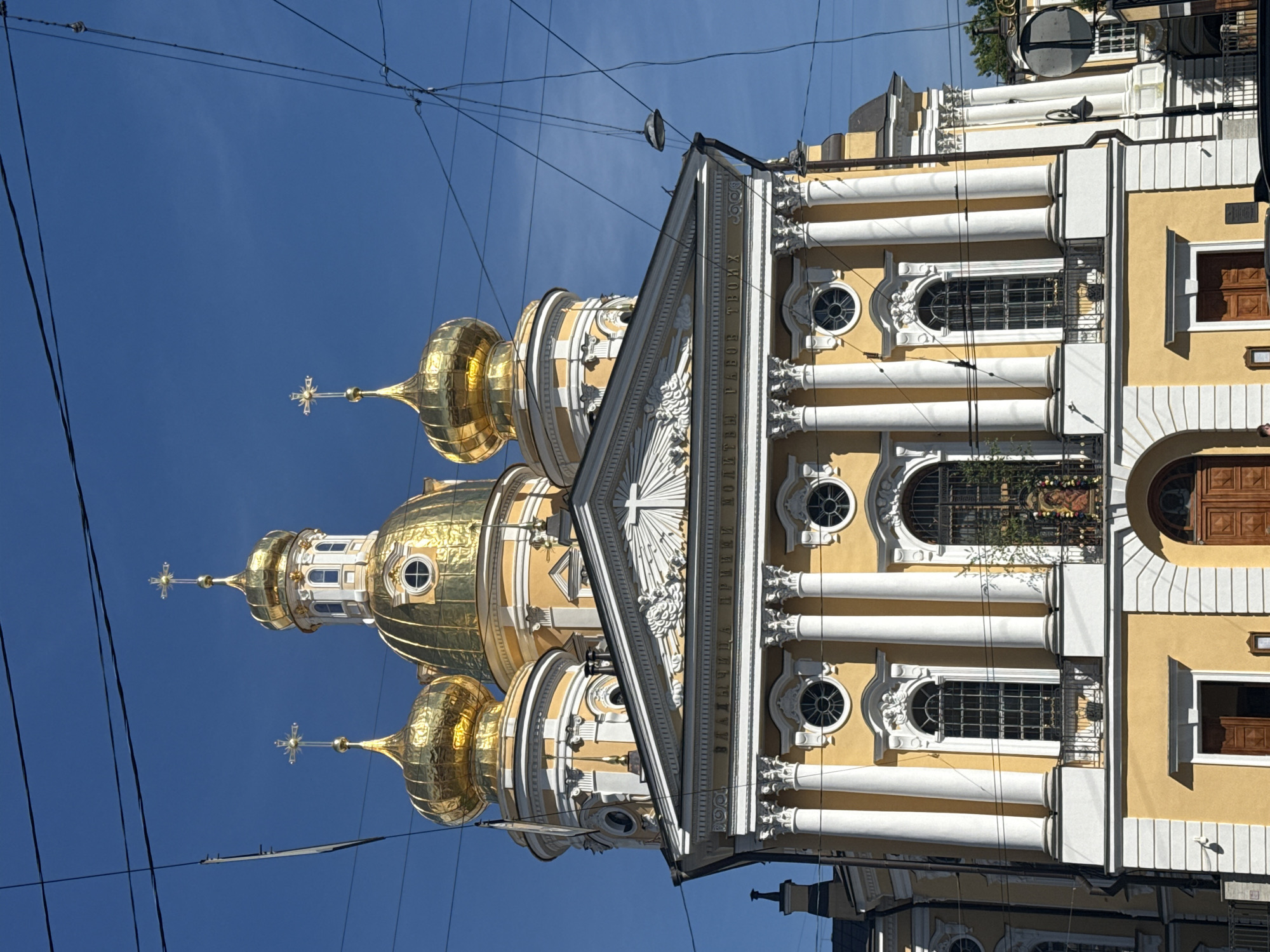 The image captures a vibrant, ornate, yellow and white Baroque-style cathedral under a clear blue sky in Saint Petersburg, Russia. The building features multiple gleaming golden onion domes topped with intricate Orthodox crosses. Four prominent golden domes are visible, with the central dome rising highest. The facade is richly decorated with white Corinthian columns, arched windows, and decorative cornices. A triangular pediment in the center, adorned with a white relief sculpture, is also visible. Below the pediment, on the frieze, Cyrillic text is discernable, reading "МОЛИТВЫ РАБОВ ТВОИХ" (meaning "the prayers of Thy servants").
The sky is bright and cloudless, indicating a clear, sunny day. Overhead, numerous dark electrical wires, possibly for trams or trolleybuses, crisscross the sky, adding a modern urban element to the historic architectural setting. No people are visible, and the scene focuses entirely on the grandeur of the cathedral itself. The bright light suggests it is midday. The architecture strongly identifies it as a Russian Orthodox church, likely a significant cathedral given its size and ornamentation.