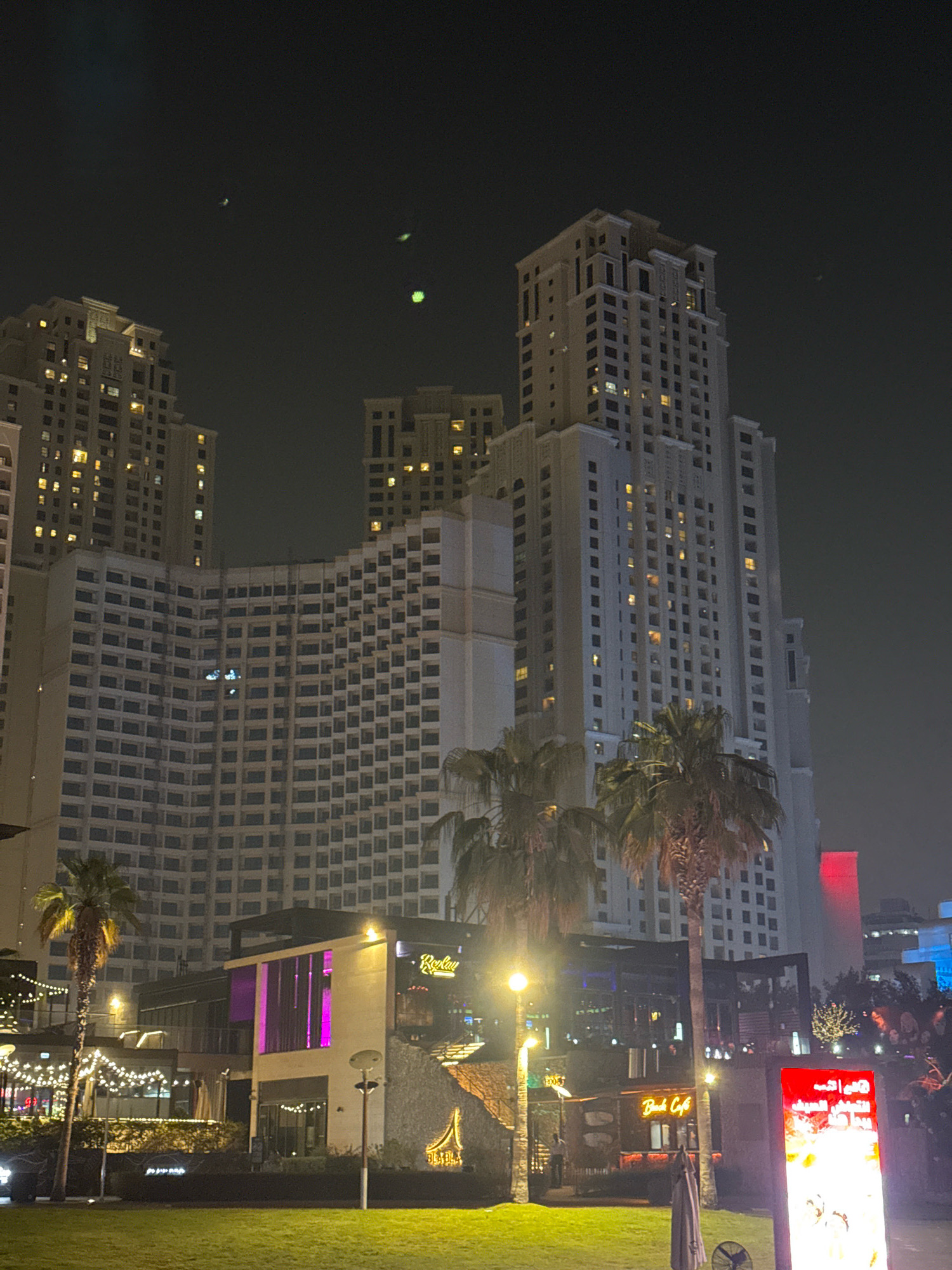The image depicts a nighttime scene in Dubai Marina, United Arab Emirates. Dominating the background are several tall, modern skyscrapers, their windows illuminated with warm yellow light, creating a stark contrast against the dark night sky. In the mid-ground, there are lower-rise buildings with illuminated sections, including one with a vibrant purple glow. Palm trees are scattered throughout the scene, adding a tropical element. In the foreground, there is a grassy area, and further back, a street or promenade with lights and some signage, including what appears to be a digital advertisement board. The overall impression is one of a lively urban environment at night, characteristic of Dubai's contemporary architecture and entertainment districts. There are no visible people or specific actions taking place.