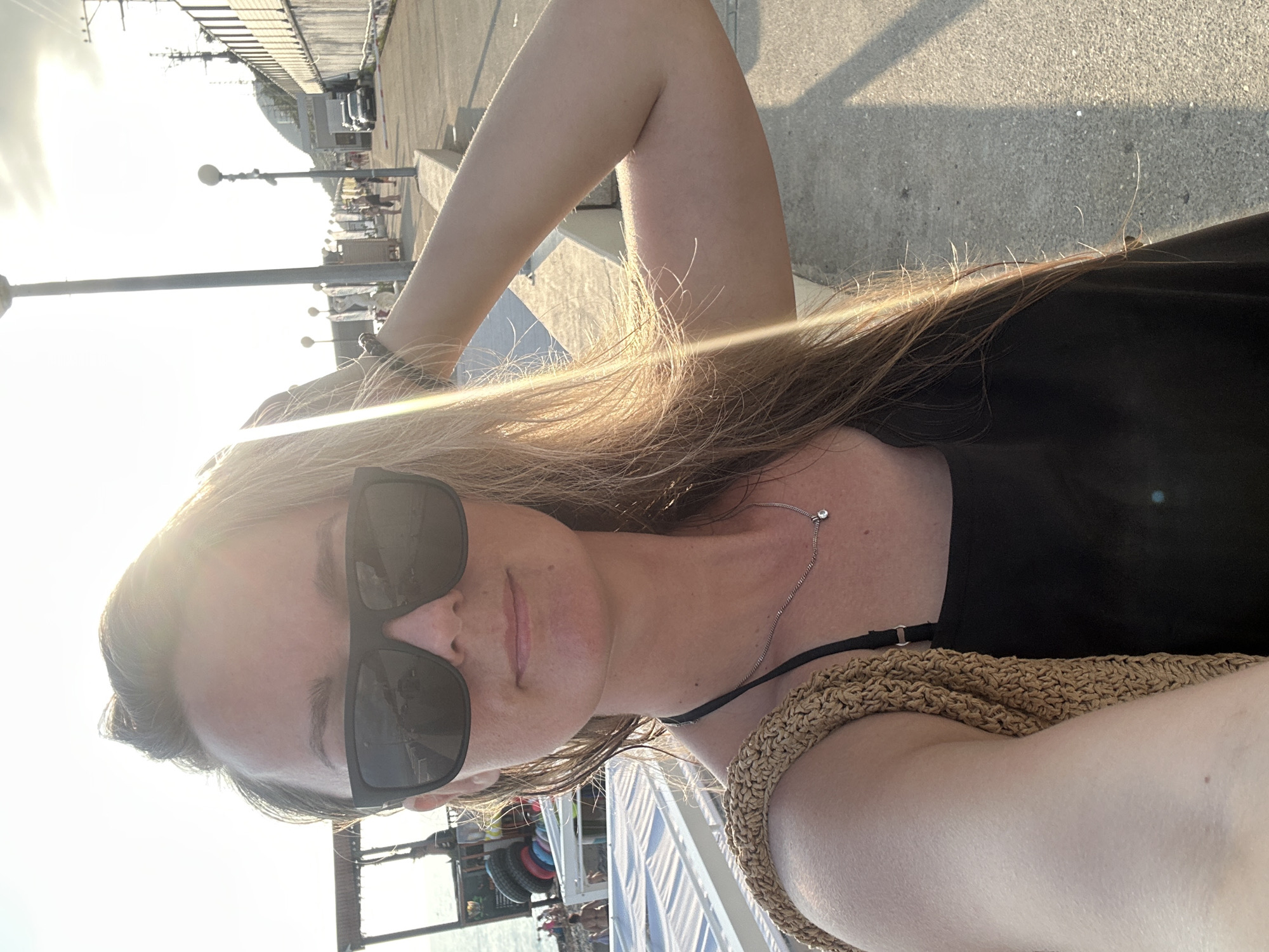 A woman wearing black sunglasses and a black tank top stands on a paved walkway near the sea. She has long, blonde hair and is wearing a straw-like bag over her left shoulder. Her right arm is raised and bent behind her head. The sun is low in the sky, casting long shadows. In the background, there are beach chairs, the sea, and a street with cars and people. The location is Krayevsko-Armyanskoye, Russia.