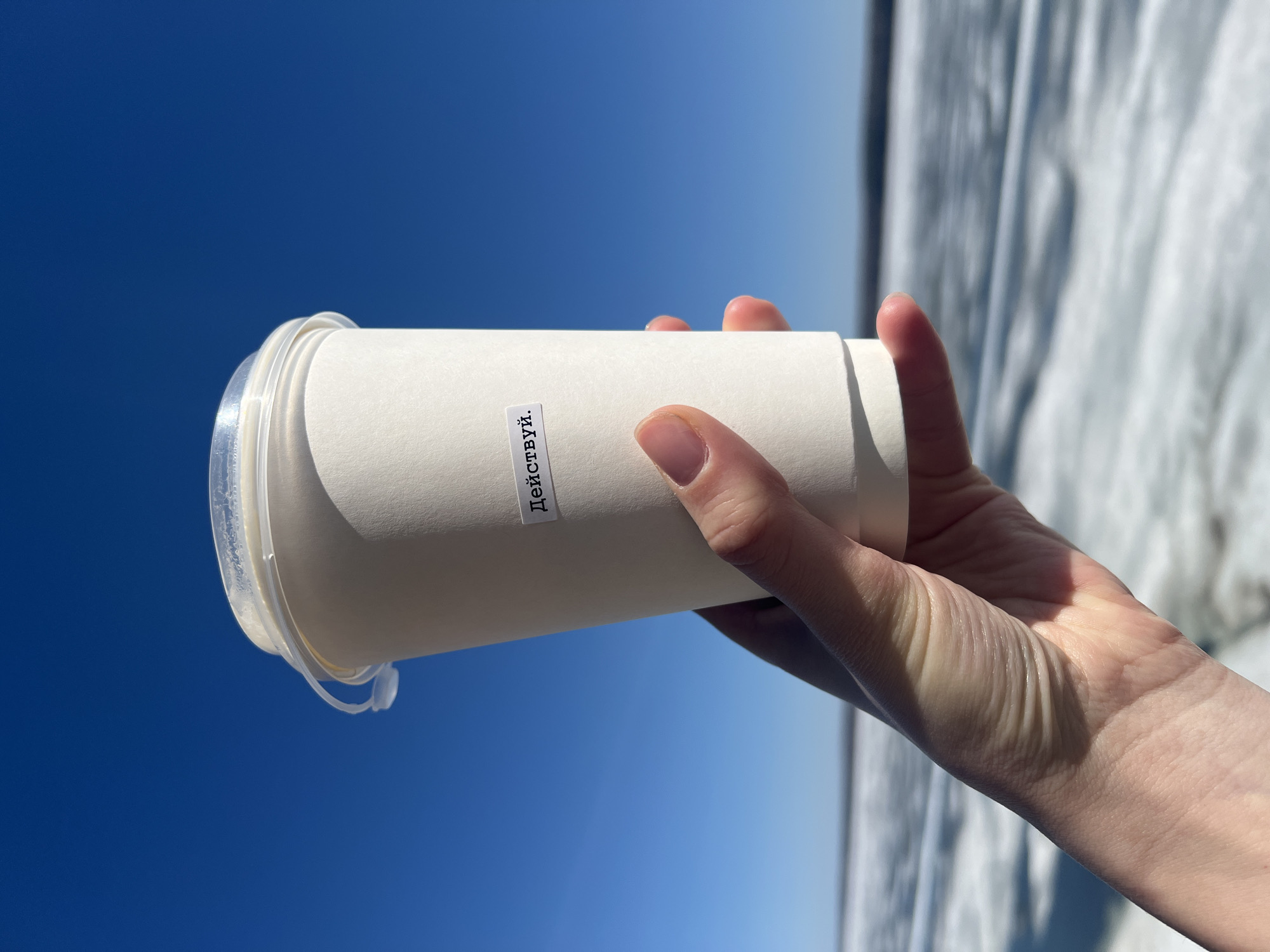 A hand holds a white, cylindrical container with a clear plastic lid against a backdrop of a clear blue sky transitioning to a light gray background. The container has a label with the Russian word "Действуй" on it. The location is unknown.