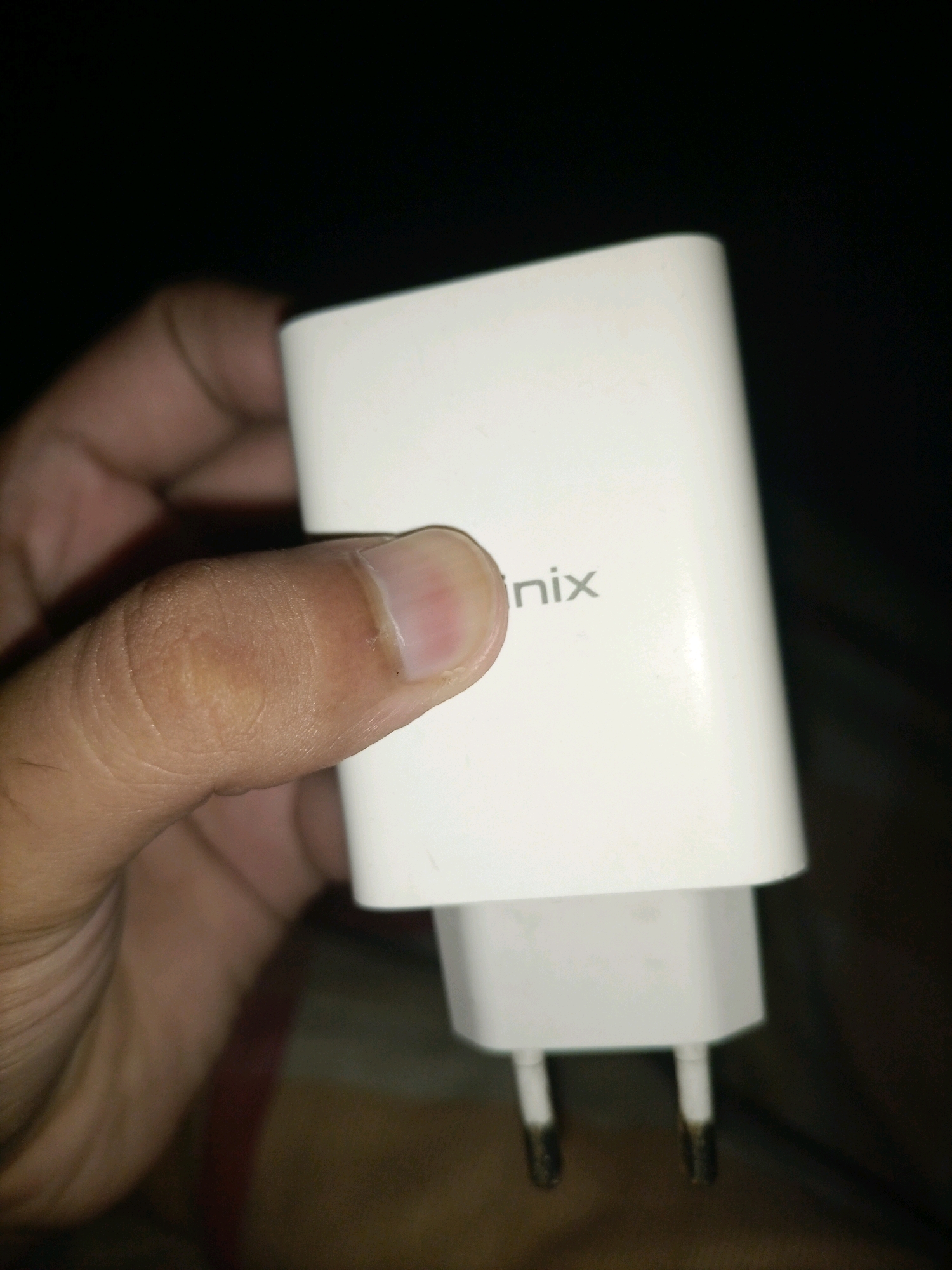 In Kangra, Pakistan, a close-up shot captures a person's thumb holding a white Infinix brand fast charger. The charger is partially obscured by the thumb, with its plug prongs visible at the bottom. The background is dark and out of focus.