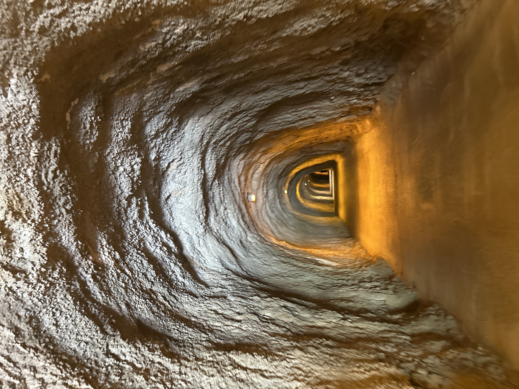 The image captures a narrow, winding tunnel or passage, appearing to descend or curve deeply into an enclosed space. The perspective looks forward and slightly downwards, following the path as it bends to the right. The walls and ceiling of the tunnel are heavily textured, resembling rough, undulating rock formations or sculpted concrete with distinct wave-like patterns and ridges. This rugged surface is illuminated by several small, embedded lights that cast a warm, yellow-orange glow, highlighting the contours and creating strong shadows. The artificial lighting suggests an interior or subterranean setting, making time of day irrelevant. The path ahead gradually darkens as it recedes into the distance, with more lights visible further along the curve. No people or visible text are present in the image. The scene conveys a sense of depth, enclosure, and a journey through a uniquely designed or natural pathway, possibly within Kamenovo, Montenegro.