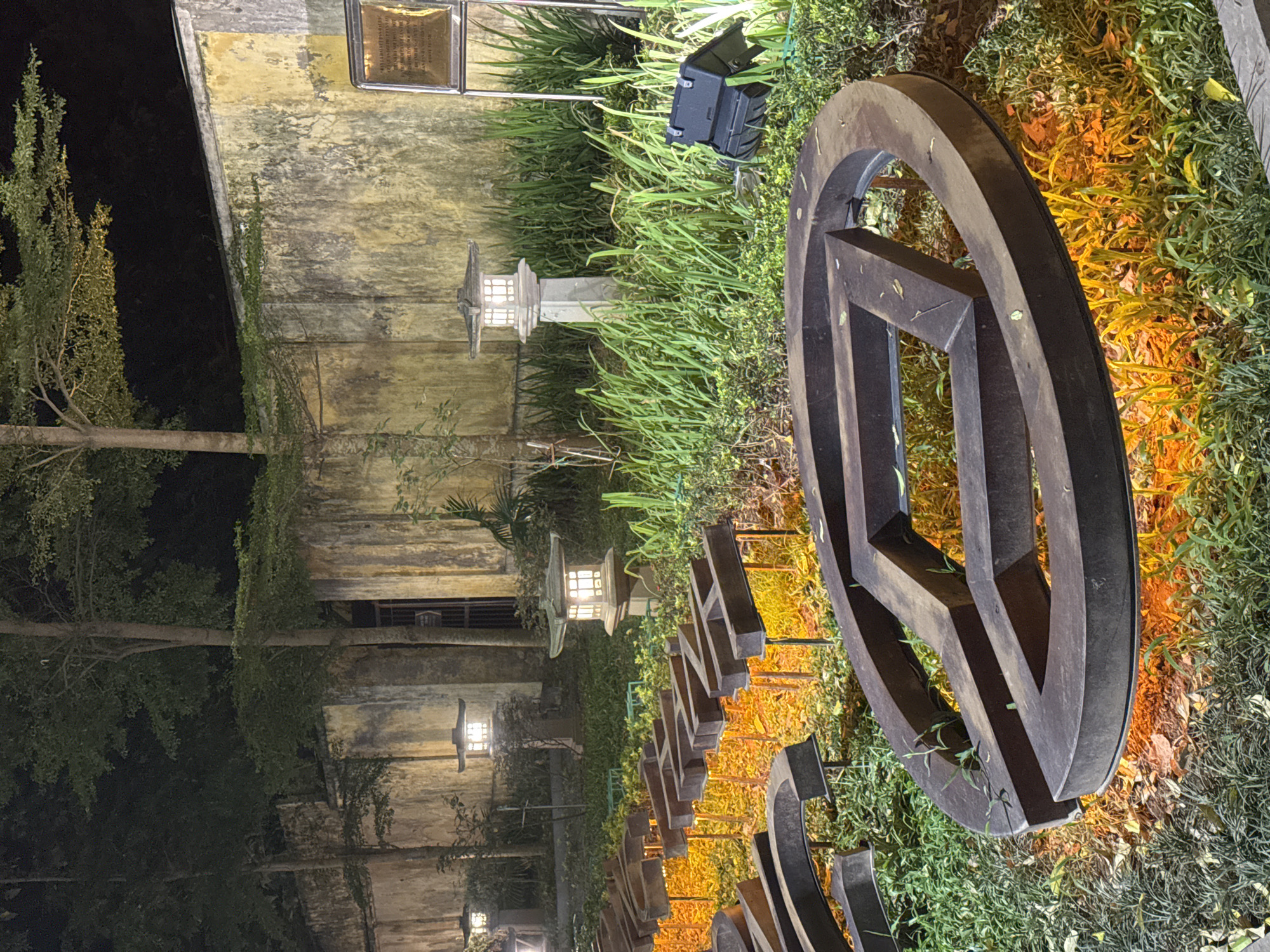 This media file, captured in Hanoi, Vietnam, depicts an outdoor scene at night, likely illuminated by artificial lighting. The foreground is dominated by a large, circular, wooden decorative element, possibly part of a sculpture or art installation. It is positioned amidst lush green foliage, which is bathed in warm, orange light, suggesting uplighting or spotlights.
Behind the wooden sculpture, several elements suggest an Asian-inspired garden or courtyard setting. Two traditional Japanese-style lanterns are visible, emitting a soft, white light. These lanterns are placed among the greenery, adding to the ambient illumination. A weathered, textured wall with a yellowish hue forms the backdrop to the left of the frame. This wall appears to have some faint markings or discoloration, adding to its aged appearance.
In the upper left portion of the image, a small, dark, box-like object is visible amongst the plants, possibly a piece of outdoor equipment or a camera. Further up on the wall, a rectangular plaque with what appears to be engraved text is affixed. The text is illegible due to the resolution and distance, but its presence suggests historical or informational significance.
The scene is characterized by a sense of tranquility and ambiance, enhanced by the interplay of light and shadow. The lush vegetation and traditional lantern designs contribute to a serene, possibly cultural, environment. The time of day is clearly night, with the dark sky and artificial lighting being prominent features. No people or specific events are captured in this static image.