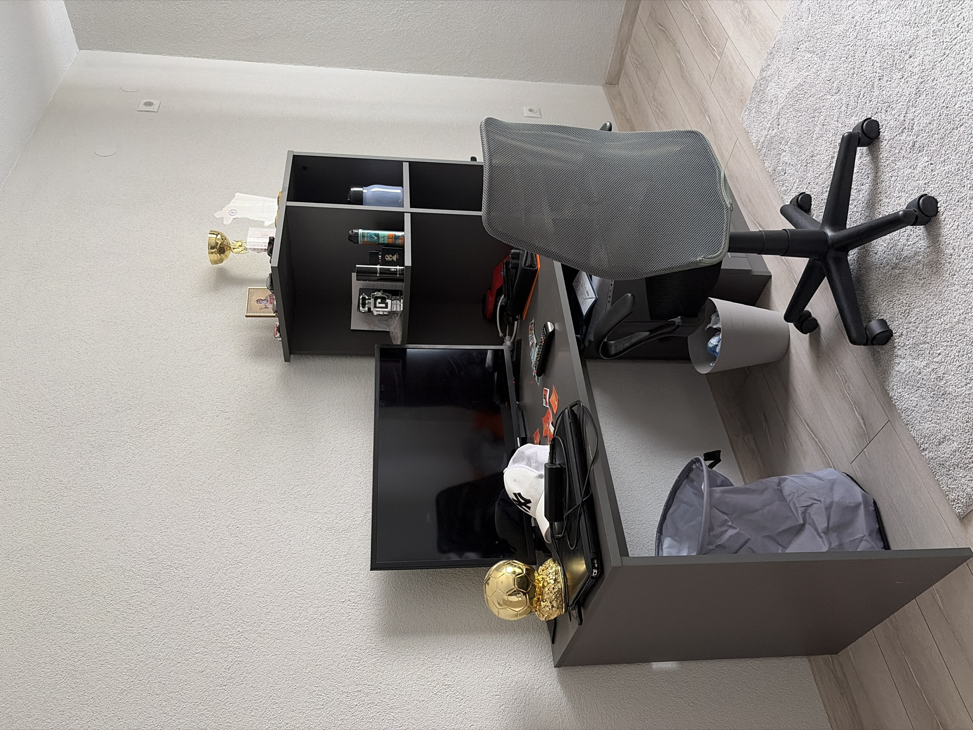 The image shows a desk with a computer monitor, a golden trophy shaped like a soccer ball, a white baseball cap, and other items. There is a black office chair in front of the desk, and a gray trash can next to the desk. The scene is set in a room with white walls and wooden floors. The location is a residential apartment in the city of Mitino, Bulgaria. There are no people visible in the image.
