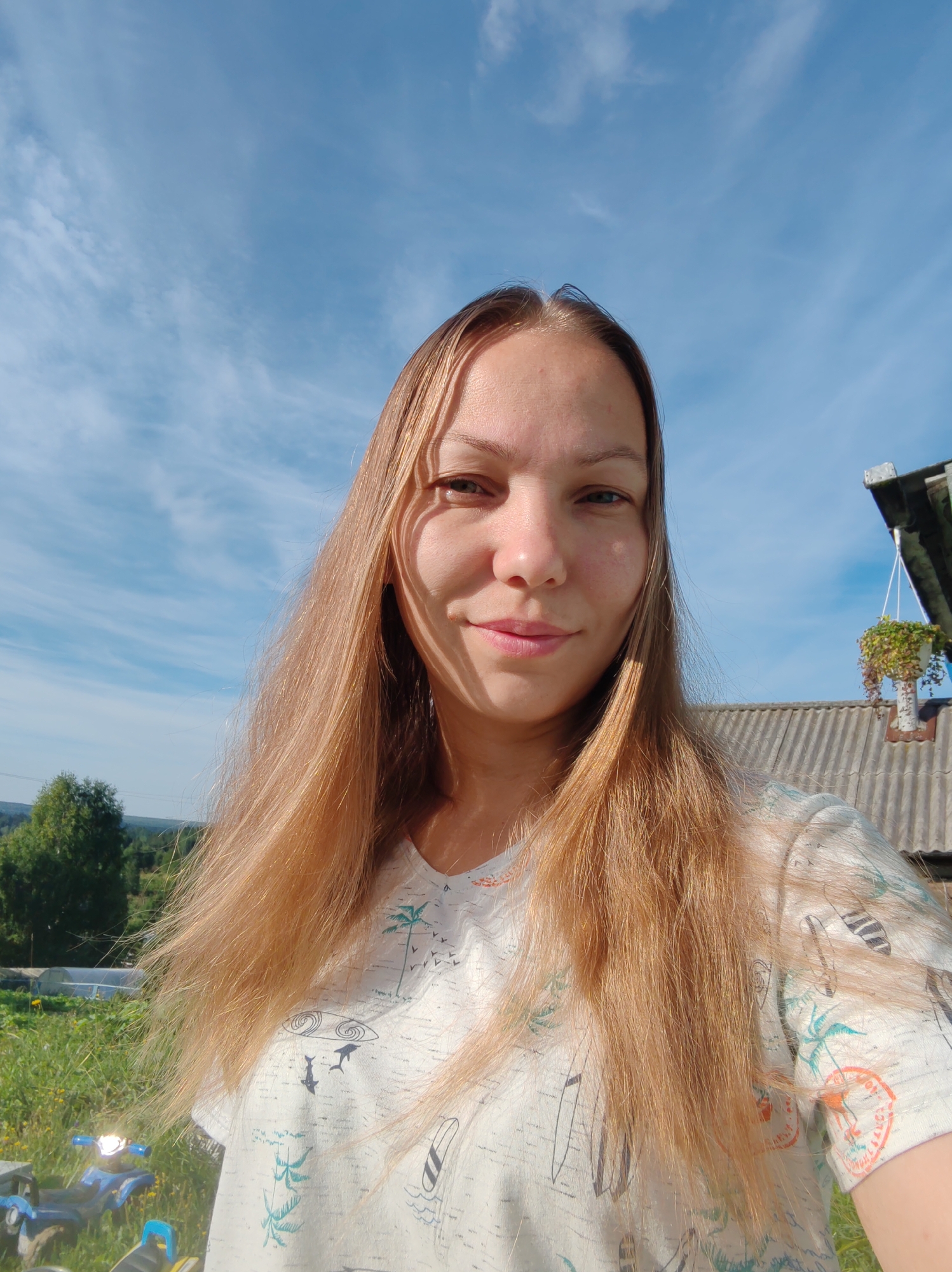 A young woman with long, blonde hair is captured in a selfie outdoors on a sunny day. She is wearing a white t-shirt with a print of palm trees and surfboards. Her face is illuminated by the sunlight, creating shadows on the left side of her face. The background features a bright blue sky with wispy clouds, a roof of a building with a hanging plant, and a green, grassy landscape with trees and possibly some small structures. The image appears to be taken in a rural setting.