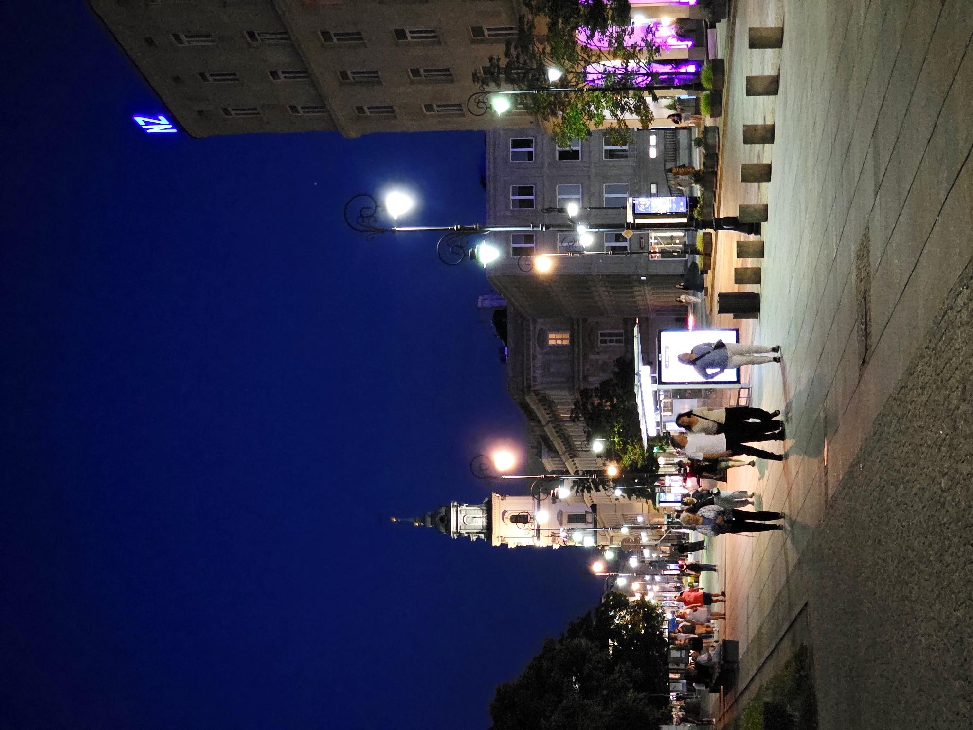 The image depicts a nighttime scene on a city street in Warsaw, Poland, likely in the Old Town or a central district. The sky is a deep blue, indicating it is twilight or early evening.
In the foreground, a wide, paved pedestrian walkway is illuminated by streetlights and ambient light from buildings. Several people are walking along the path. Some are walking in pairs, while others are alone or in small groups. They are dressed in casual attire, consistent with a warm evening.
To the left, a row of trees lines the walkway, casting shadows. Further down the street, a prominent church tower with a spire rises against the night sky, its architecture suggesting historical significance. Streetlights with ornate designs illuminate the pathway.
On the right side of the image, a multi-story building with many windows is visible. A neon blue sign displaying "NZ" is prominently placed at the top of this building. In front of this building, there are trees with purple lighting, adding to the vibrant nighttime atmosphere. Other buildings, appearing to be of similar age and style, line the street.
A bus stop shelter with glass panels is also visible, and several concrete bollards are spaced along the edge of the walkway. The overall impression is a well-lit urban environment, alive with pedestrian activity at night.
