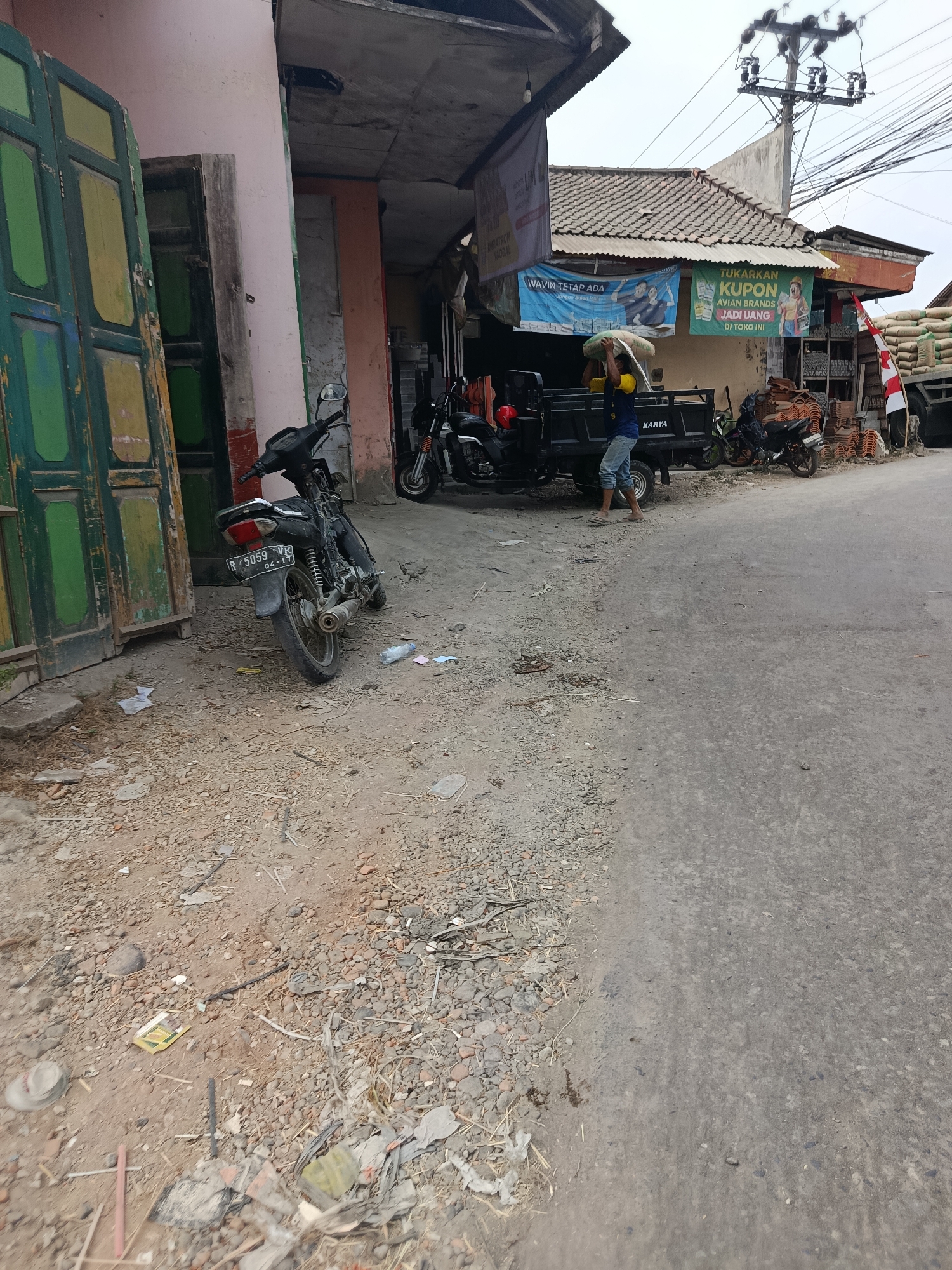 The image depicts a street scene in Rawajaya, Indonesia. A black motorcycle is parked on the left side of a dirt and gravel road. Adjacent to the motorcycle is a building with a pink facade and a large, weathered wooden door painted in green and yellow. Further down the street, a black utility truck is parked, and a person wearing a blue shirt and jeans is carrying a sack of grain over their shoulder, seemingly unloading it from the truck. A green banner with Indonesian text and an image of people is hanging above the truck. In the background, several other motorcycles are visible, as well as stacks of roof tiles and bags of materials, suggesting a commercial area, possibly a warehouse or market. Electrical poles and wires are present overhead, and the sky is overcast. The overall environment appears somewhat unkempt, with debris scattered on the ground.