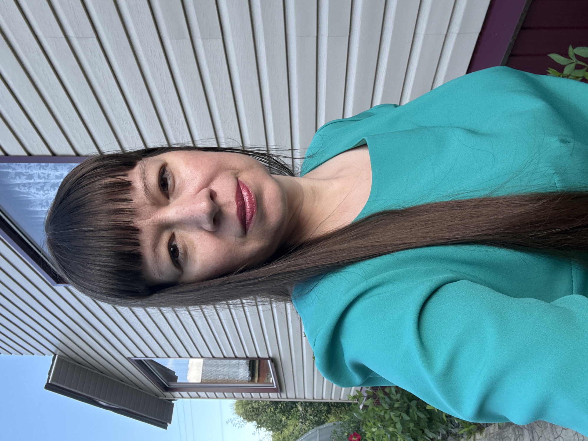 A woman with shoulder-length brown hair and bangs is pictured in a close-up, medium shot, facing the camera. She is wearing a teal-colored dress with a square neckline and puffed sleeves. The background consists of a light-colored, horizontally-paneled house siding and a clear blue sky. To the left of the woman, there is greenery and a hint of a red rose. The overall scene suggests an outdoor setting, possibly in a residential area of Vetluzhsky, Russia.