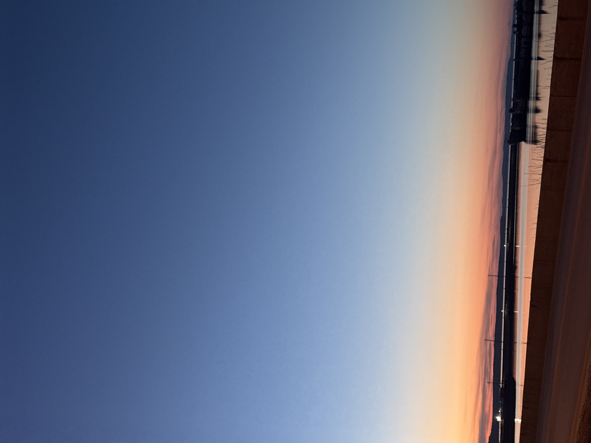 The image shows a horizontal view of a gradient sky, transitioning from deep blue to a pale orange near the horizon. A strip of land or buildings is visible along the right side of the image, appearing dark against the brighter sky, possibly in Chaikovsky, Russia.