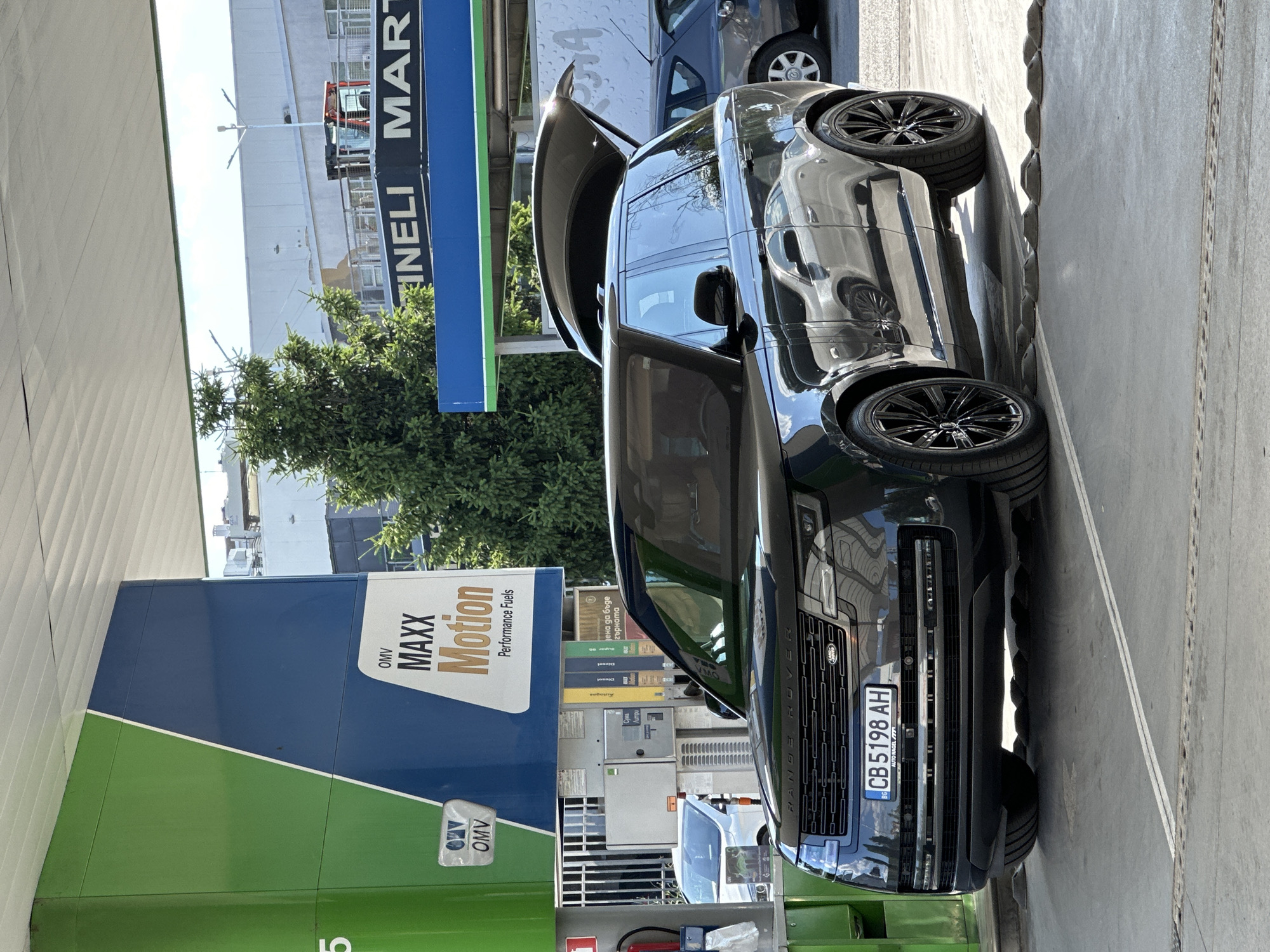A black Range Rover is parked at an OMV gas station in Veliko Tarnovo, Bulgaria. The car's tailgate is open, and the front of the vehicle is visible. The gas station has green and blue signage with "OMV MAXX Motion Performance Fuels" printed on it. Other cars and a tree are also visible in the background. The scene appears to be taking place during the daytime, under sunny skies. The license plate of the Range Rover reads "CB 5198 AH".