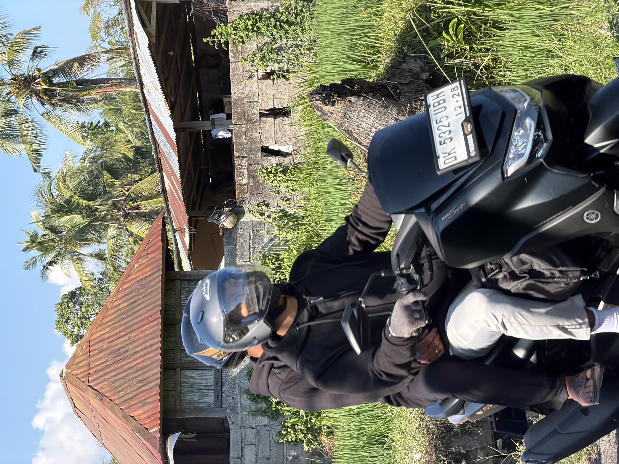 Two individuals are riding a black motorcycle on a paved road in Sawan, Indonesia. The person in front, wearing a black hoodie and grey gloves, is operating the motorcycle. They have a gray helmet with a clear visor. The person behind, also wearing a black hoodie, has their arm around the driver. They are wearing a helmet with an orange and black design. The motorcycle has a license plate that reads "DK 5325 UBH 12-28".
The environment is rural and tropical. To the left of the road, there is a rustic building with a corrugated metal roof. Behind the building, there are palm trees and lush green vegetation. To the right of the road, there are vibrant green rice paddies. A weathered tree trunk is visible in the foreground to the right. The sky is clear blue with a few scattered clouds. The scene appears to be captured during daylight.