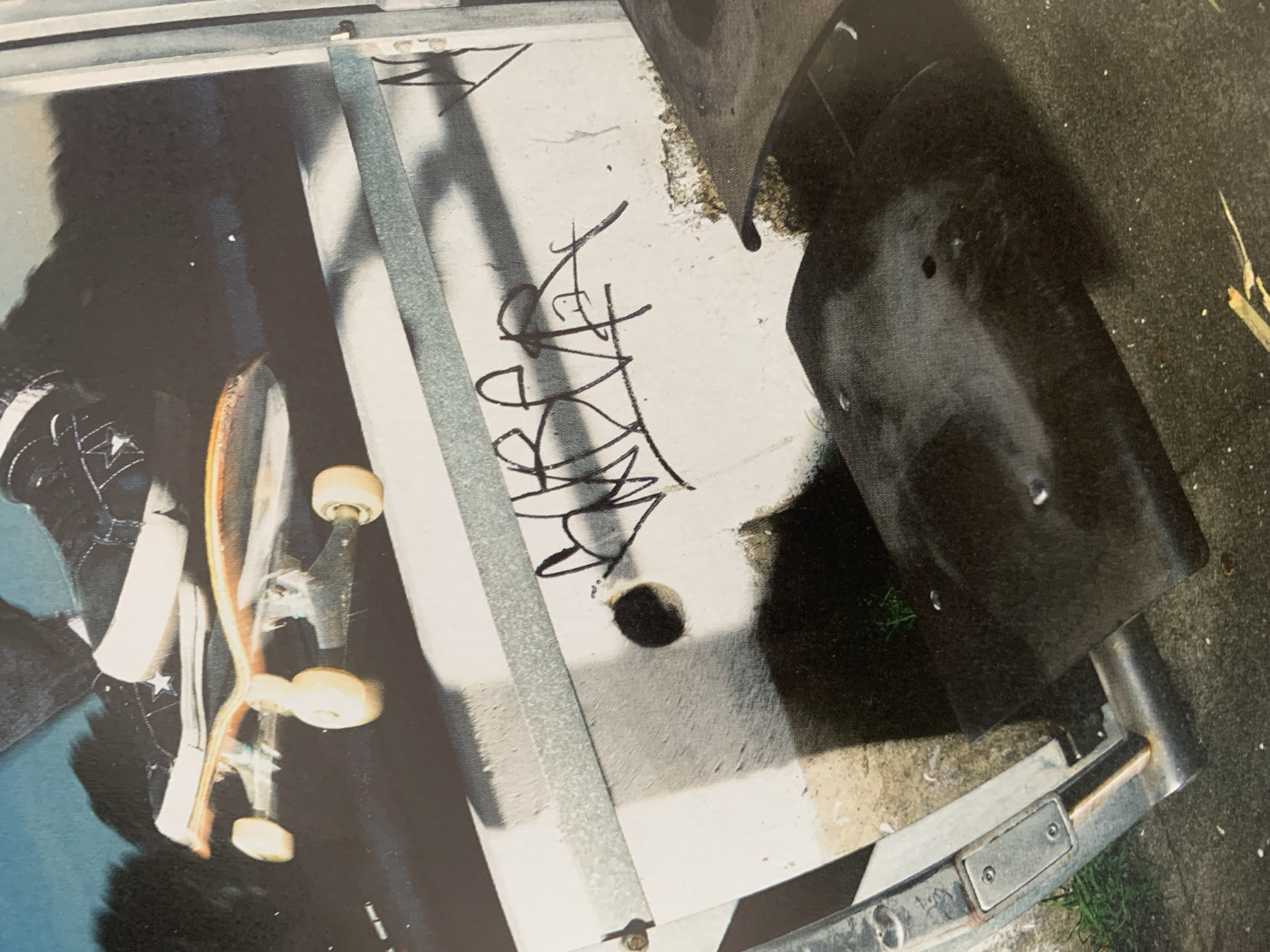 A skateboard is partially visible on a surface next to a curb with graffiti. The environment appears to be an urban street setting, possibly in Barcelona, Spain. A foot in a black and white Converse shoe is also visible in the image. The skateboard is positioned near the edge of what seems to be a metallic structure. The curb features black graffiti. A black object obscures part of the curb. The composition includes concrete and metal elements, suggesting an urban landscape.