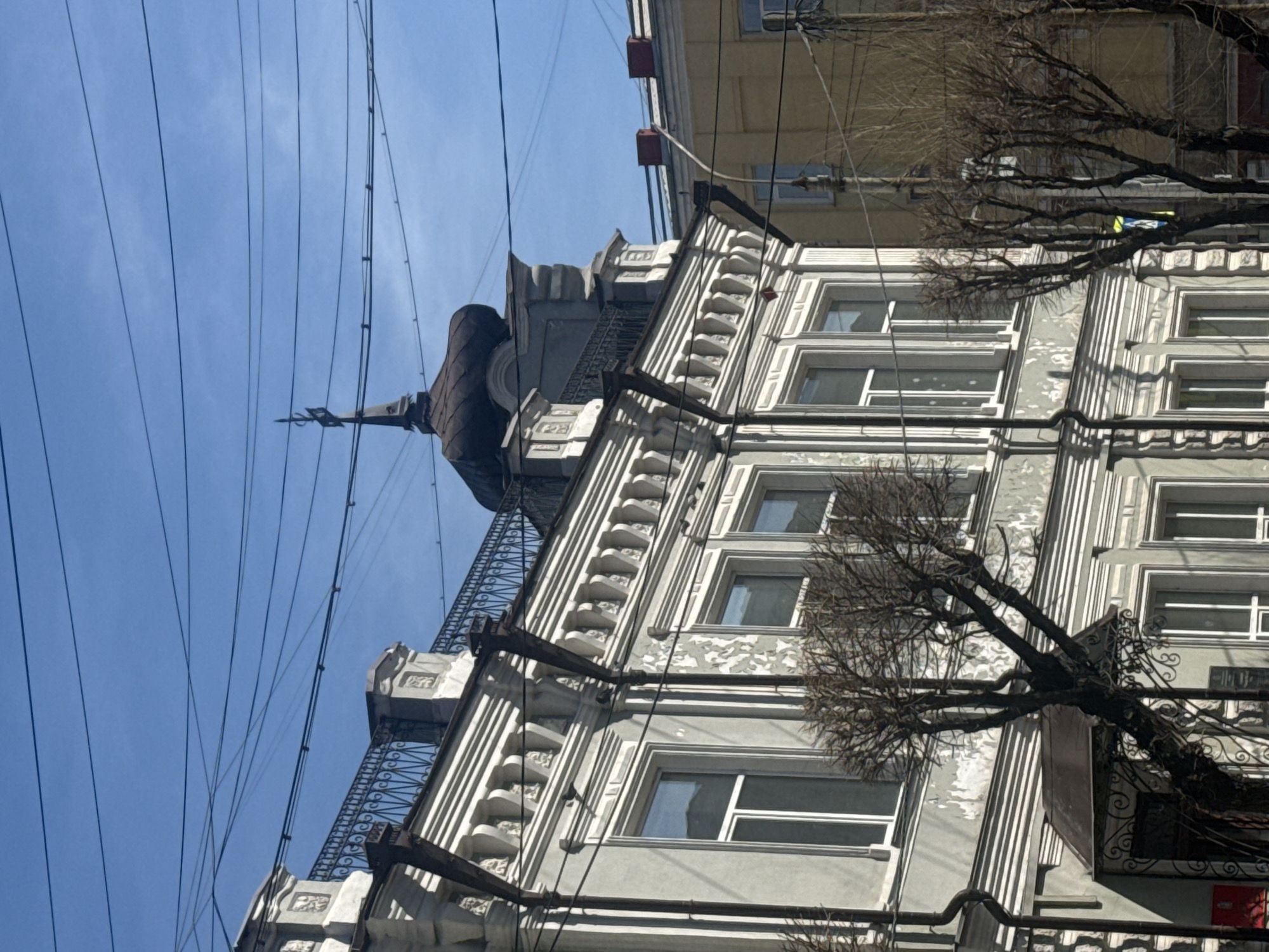 The image displays an upward-looking view of an ornate, light-colored building with a clear blue sky in the background, crisscrossed by numerous overhead wires. The primary building, which appears to be grey or off-white, features a richly decorated facade with vertical fluting, elaborate window frames, and decorative elements resembling cornices and balustrades. Wrought-iron railings are visible on what appear to be balconies or decorative ledges.
Towards the upper left side of this prominent building, a distinctive dark, conical turret with a pointed spire or weather vane stands out against the sky. The building's exterior shows signs of age, with some areas of worn or peeling paint.
In the background, behind the main structure, parts of a simpler, yellow or beige building can be seen. Bare, gnarled tree branches, devoid of leaves, are prominent on the right side of the image, extending into the frame and partially obscuring the building, suggesting a late winter or early spring season.
The sky is a bright, clear blue, indicating sunny weather during daytime. There are no people visible, and no specific activity is taking place. The dense network of overhead wires suggests an urban setting. The architecture, with its detailed ornamentation and turret, is characteristic of older city buildings, aligning with the provided context of Krasnoyarsk, Russia. There is no visible text.