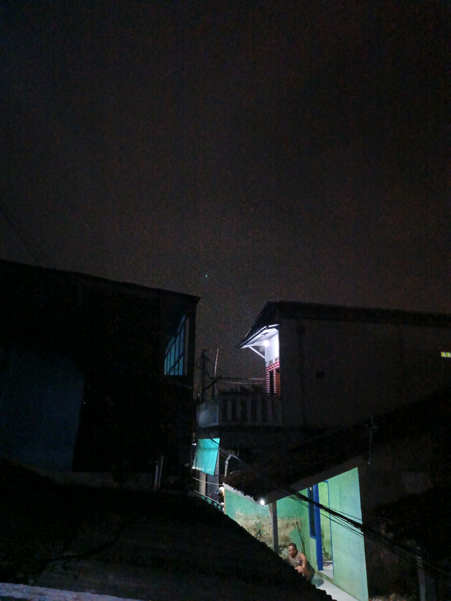This is a nighttime exterior shot of a residential area in Dayeuhkolot, Indonesia. The image is dark, with only artificial lights illuminating parts of the scene. Two buildings are visible, appearing to be houses or small structures. One building on the left is mostly in shadow, with a sliver of light coming from a window. The other building on the right has a brightly lit entrance area. A person is partially visible sitting on the ground in front of the building on the right, illuminated by its light. Power lines are strung across the scene, and parts of rooftops are visible in the foreground. The sky above is dark and cloudy.