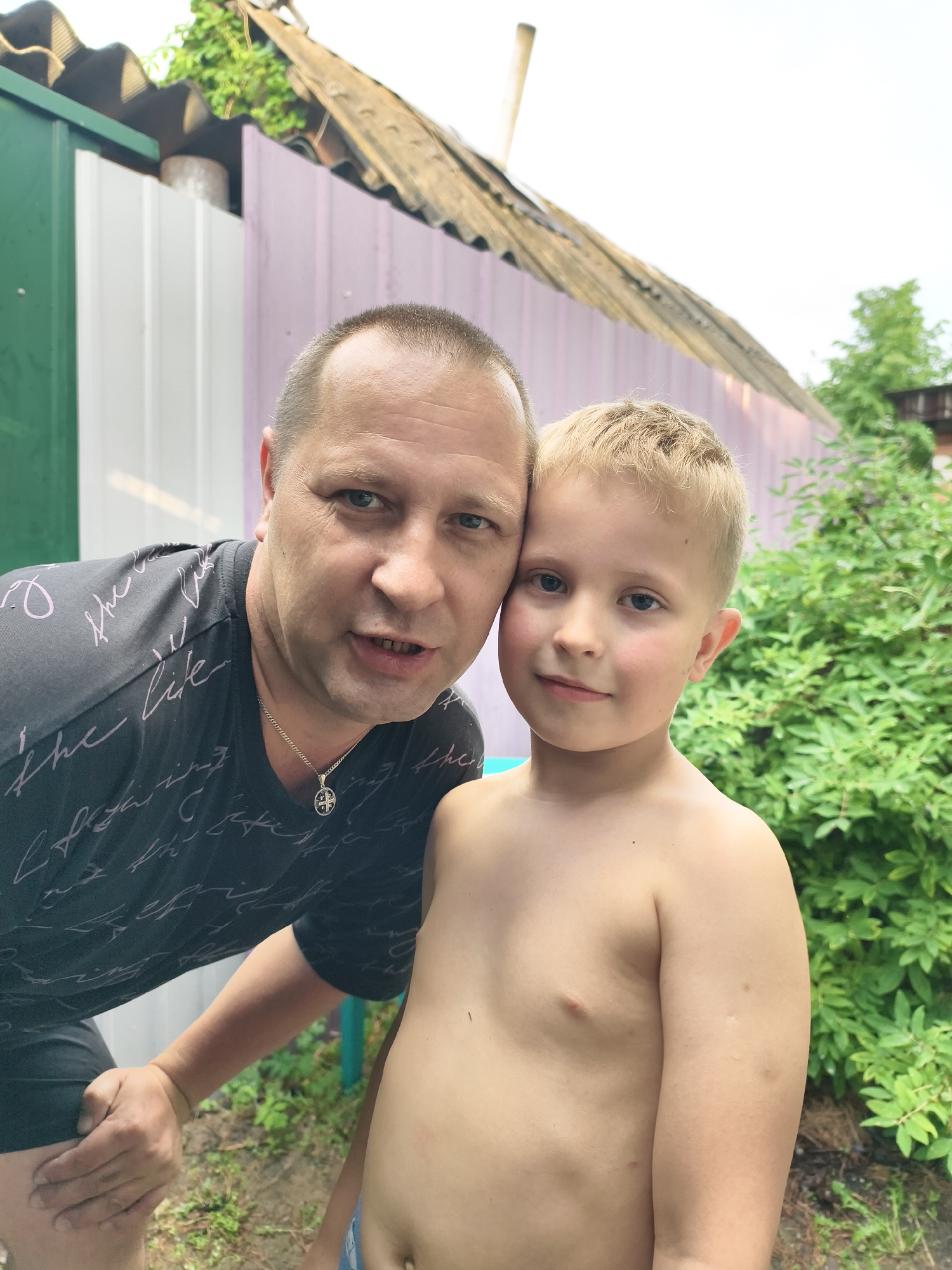 This is a close-up, waist-up portrait of a man and a young boy. The man, on the left, is bald and has a fair complexion. He is wearing a dark t-shirt with a repeating pattern of light-colored cursive text. Around his neck, he wears a silver chain with a circular pendant. His left hand is resting on his thigh, and he is leaning towards the boy. His facial expression suggests he is speaking or interacting with the person taking the photo.
The boy, on the right, has blond hair and fair skin. He is shirtless and looking towards the camera with a gentle smile. His arm is resting at his side. The man and boy are standing close together, with their heads nearly touching.
The background shows an outdoor scene. Behind them is a fence with sections of green, white, and light purple. Above the fence, a rustic, wooden roof structure is visible, possibly part of a shed or outbuilding. Green foliage from bushes or trees is present on the right side of the frame. The lighting suggests it is daytime, likely overcast given the soft light. The overall mood appears to be casual and affectionate, capturing a moment between the two individuals. There is no clear indication of the specific time of day or weather conditions beyond the general lighting.