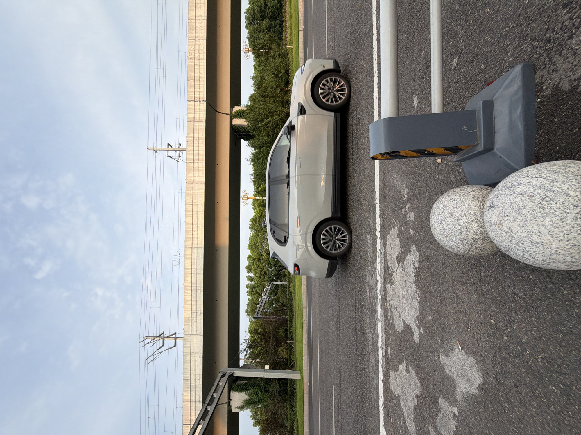 The image depicts a light-colored SUV driving on a paved road in Jiangjin, China. The car is in motion, captured in a side profile view. The road has visible white lane markings. In the foreground, a gray traffic barrier with yellow and black reflective strips is prominent, situated next to two round, speckled bollards. The background features a raised concrete structure, likely an overpass, with electrical wires and poles running above it. A dense line of green trees forms the horizon behind the road. The sky is partly cloudy with a pale blue hue, suggesting either early morning or late afternoon. The scene is set outdoors in an urban or semi-urban environment.