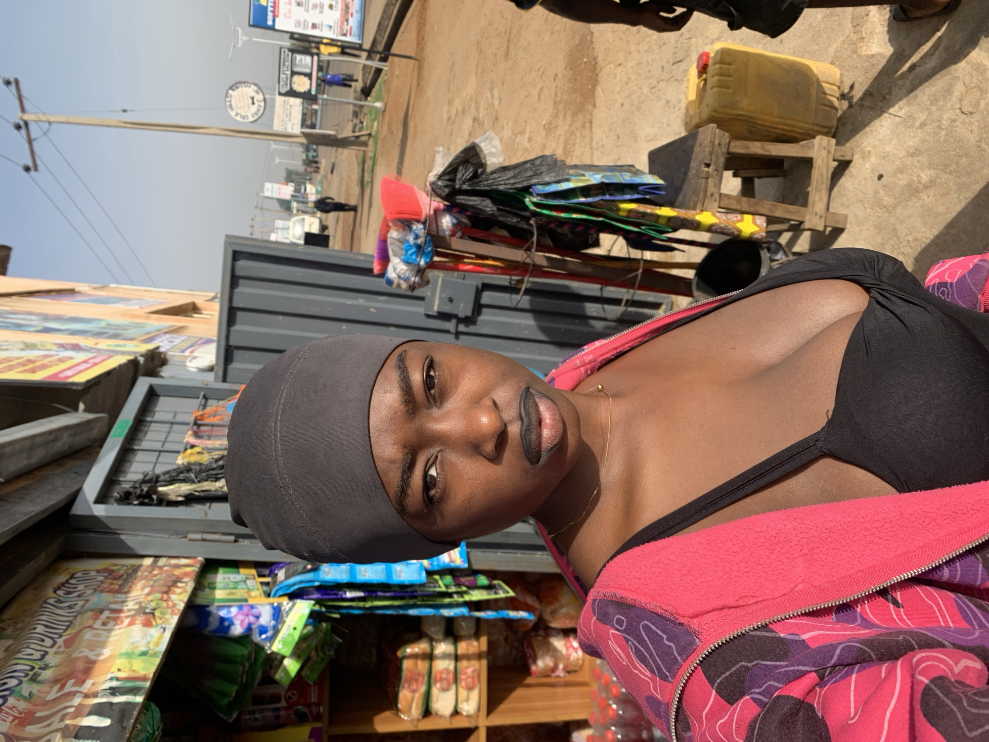 A woman with dark skin and a gray head covering poses for the camera. She wears a black top and a pink patterned jacket. She stands in front of a small street vendor stand with various goods on display. The stand is made of metal and wood and holds snacks, candy, and other items. The surrounding environment shows a city street with utility poles, signs, and distant buildings. Based on the prompt, this scene is situated in Jos, Nigeria. Other items include a yellow jerrycan on a stool, brooms, a black bucket, and advertising posters on the vendor stand.