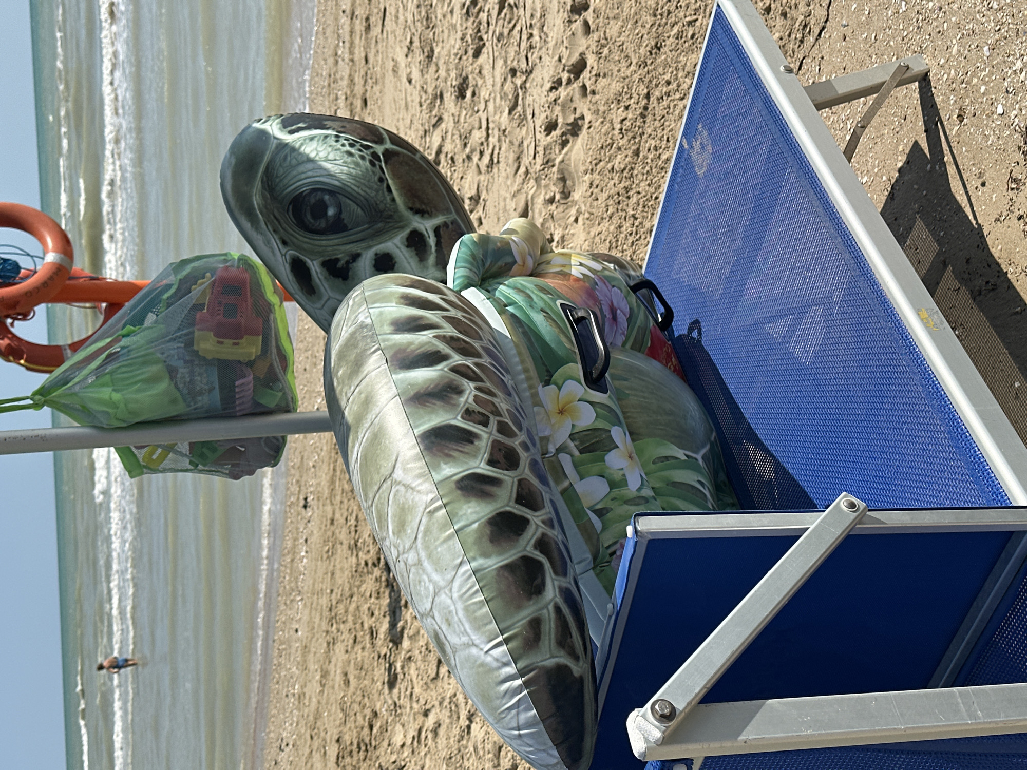 An inflatable turtle float rests on a blue beach chair on a sandy beach. Behind the float, a green mesh bag filled with toys hangs from a pole, and a life preserver is visible. The ocean with gentle waves is in the background, and a person is standing in the water. The scene is likely located in Pineto, Italy, a coastal town known for its beaches.