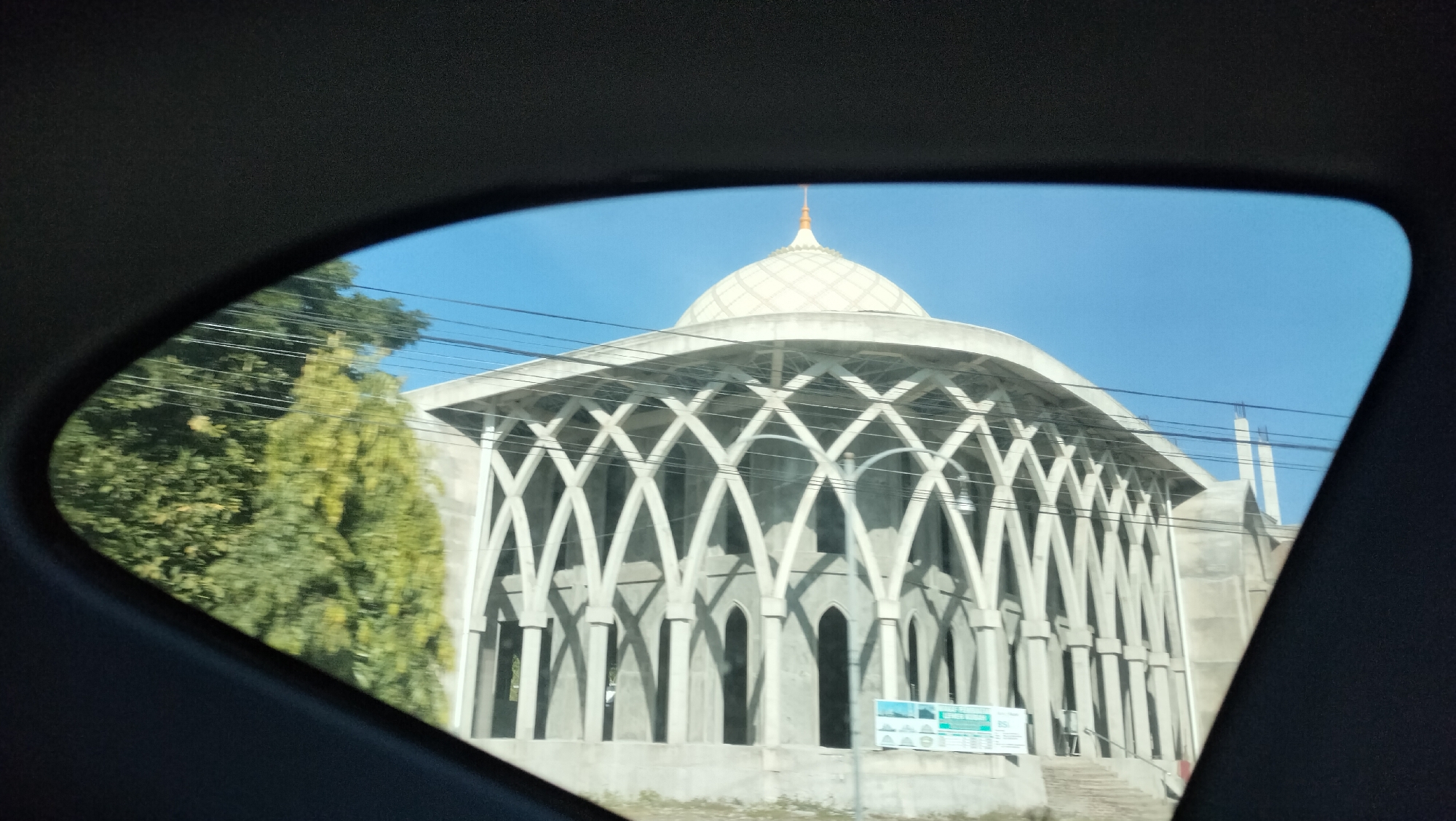 The image, taken from inside a vehicle looking through a window, shows the exterior of a mosque under construction in Dayah Caleue, Indonesia. The mosque features a large, white dome with a pointed finial, and a distinctive facade with a series of repeating arched columns creating a lattice-like pattern. The structure appears to be made of concrete and is unfinished, with scaffolding visible in some areas. To the left of the mosque, a lush green tree with yellowing leaves is partially visible. Overhead, power lines crisscross the clear blue sky. A sign with text and logos is positioned in front of the mosque's steps. The overall scene depicts a construction site against a bright, sunny day.