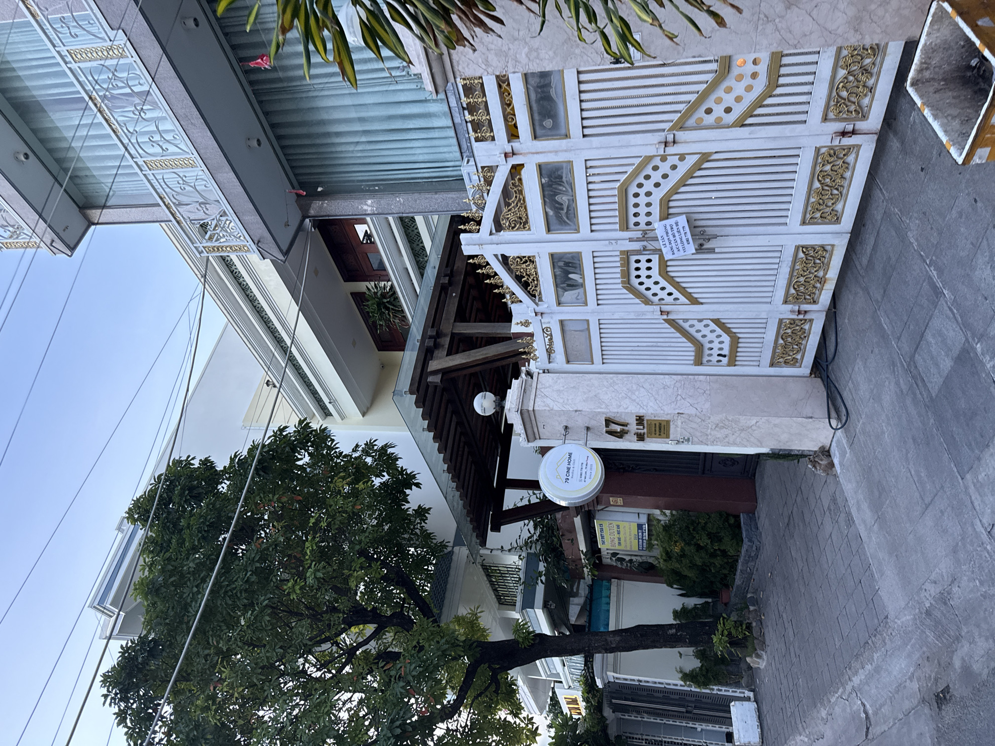 The image depicts the entrance to a residential property on a paved street in Nha Trang, Vietnam. A large, ornate white and gold metal gate stands prominently in the foreground, partially open. A white sign with black text reading "LETAN BANG DON PHONG AC CAN HO TRO VE LONG LIEN HE: 0382 778 779" is attached to the left gate panel. To the right of the gate, a light-colored stone pillar bears the numbers "47" in black. Above the gate and pillar, a modern multi-story building with large windows and white balconies is visible, with electrical wires running across the frame. A mature tree with green foliage stands to the left of the gate, casting shadows on the sidewalk. The sidewalk is paved with grey tiles, and a metal trash bin with yellow and white markings is situated on the right side of the image. The overall scene suggests a residential street in a well-maintained urban area.