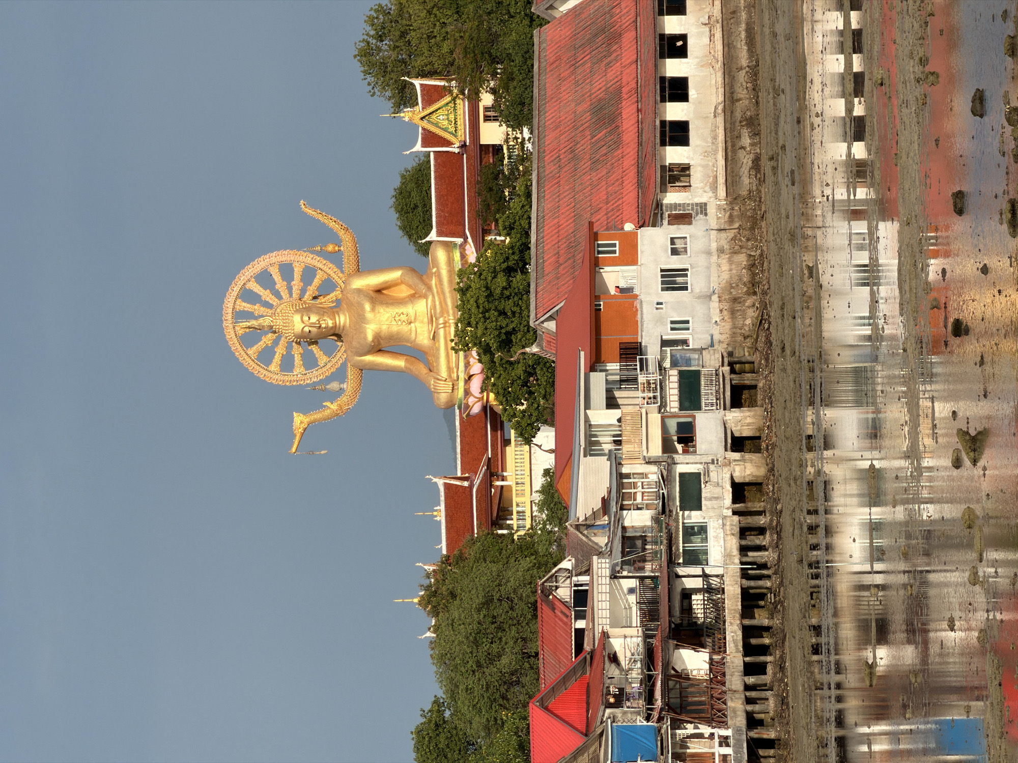 The image depicts the Big Buddha statue of Wat Phra Yai in Ko Samui District, Thailand, situated on a hillside overlooking the coast. In the foreground, a row of stilt houses with colorful roofs line the waterfront. The water level appears low, revealing mudflats and rocks. The Big Buddha, a prominent golden statue with a radiating halo, is visible in the background, behind several tiered temple buildings with red roofs and ornate golden spires. The sky above is a clear, pale blue. The overall scene is a mix of religious architecture and everyday coastal life.