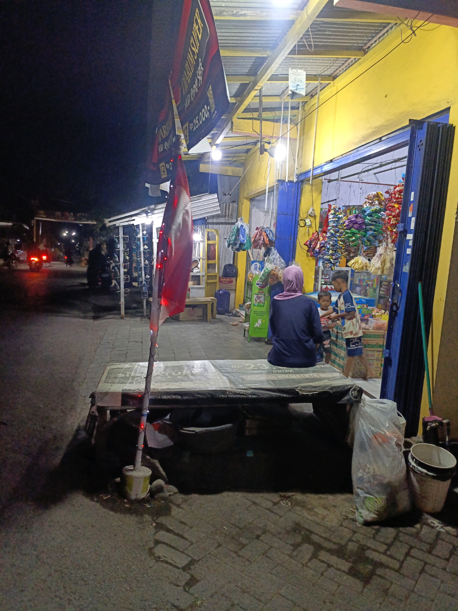 A night-time scene unfolds in Betek, Indonesia, at a roadside store. A woman wearing a purple hijab and a dark blue top is seated facing away from the camera, looking towards the store entrance. Two young boys are interacting with her. The store is brightly lit, showcasing shelves stocked with various colorful snack bags and products. One boy, wearing a t-shirt with the number "25" prominently displayed, is handing something to the other boy. In the foreground, a low-lying table with a patterned surface is visible, with a flag pole adorned with red lights standing beside it. To the right of the table, a large plastic bag filled with assorted trash sits next to a white trash can. The street in front of the store is paved with cobblestones, and a few vehicles, their taillights illuminated, are visible in the background, suggesting active street traffic.