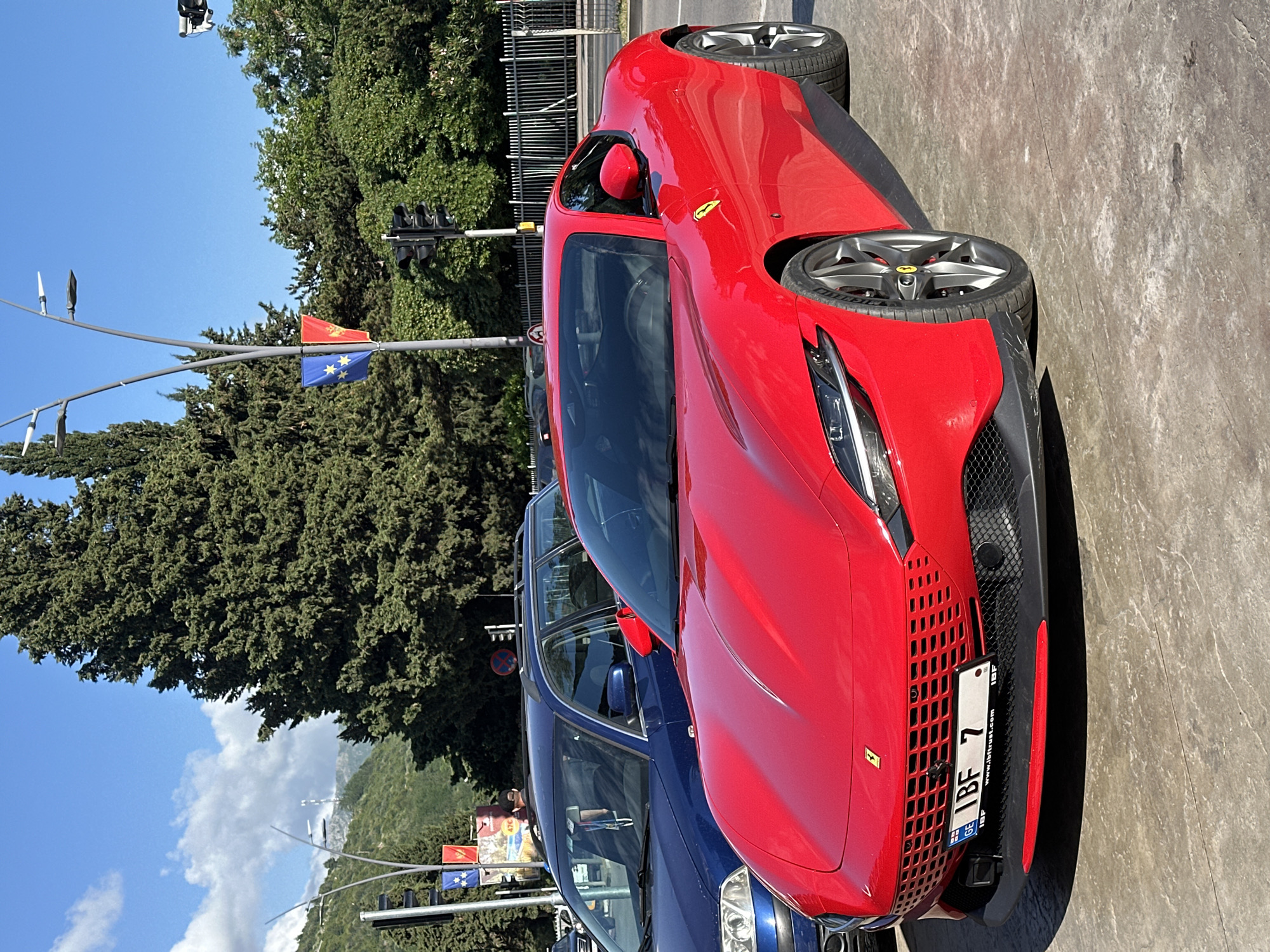 The image depicts a red Ferrari parked on a street in Budva, Montenegro. The vehicle is positioned next to a blue car. Traffic lights and flag poles are visible nearby. The flags consist of a Montenegrin flag and a European Union flag. A large green tree dominates the left side of the frame. The sky is blue with clouds in the distance.