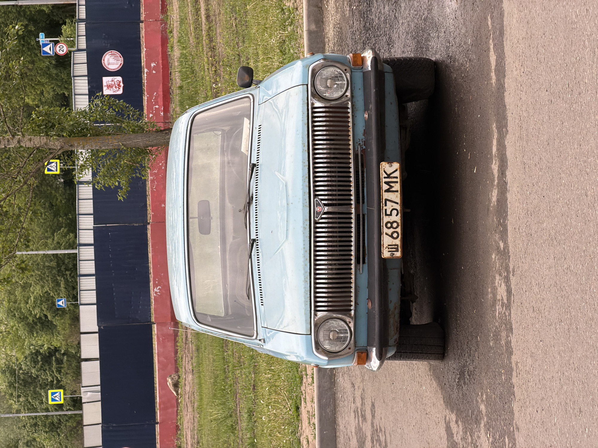 The image shows the front of a light blue GAZ-24 Volga car parked on a street. The car has a Soviet-era license plate that reads "Ш 68-57 MK." The car is parked next to a curb and sidewalk, with grass visible between the car and a blue and red fence. The fence has various road signs and circular emblems attached to it. A tree partially obscures the fence and road signs. The location is Moscow, Russia, as indicated by the license plate style. The street surface appears to be asphalt.