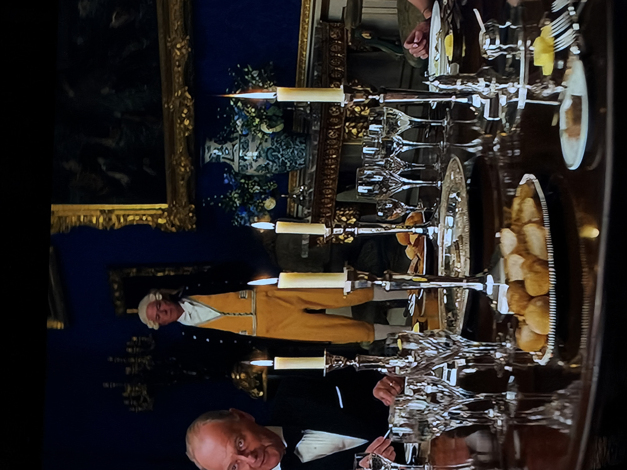 The image captures a lavish and formal dinner scene, likely set in a grand dining room during the evening or night, illuminated primarily by candlelight.
In the foreground, an older man, appearing to be actor Anthony Hopkins, is seated at a table, looking directly at the viewer with a serious or intense expression. He wears formal evening attire, including a black tuxedo with a white shirt and bowtie. His hands are visible near silverware on the table.
The large dining table is elaborately set with multiple ornate silver candelabras holding lit candles, creating a warm, glowing atmosphere. Numerous clear glass goblets and wine glasses of various shapes are arranged on the table. Silver platters laden with what appear to be bread rolls or pastries, along with white plates and silverware, are also visible.
In the mid-ground, standing to the left of the table, is another man dressed in historical period attire. He wears a white powdered wig, a striking mustard yellow or gold-colored coat, a white shirt, and breeches. He appears to be observing or participating in the event.
The background features a dark blue wall adorned with a large, golden-framed painting, which is too dark to discern its subject. Further back, a decorative vase filled with blue and white flowers adds to the opulent setting. The overall scene suggests a high-society or historical banquet, characterized by its rich decorations, formal dress, and elegant candlelight. There is no visible text in the image.
