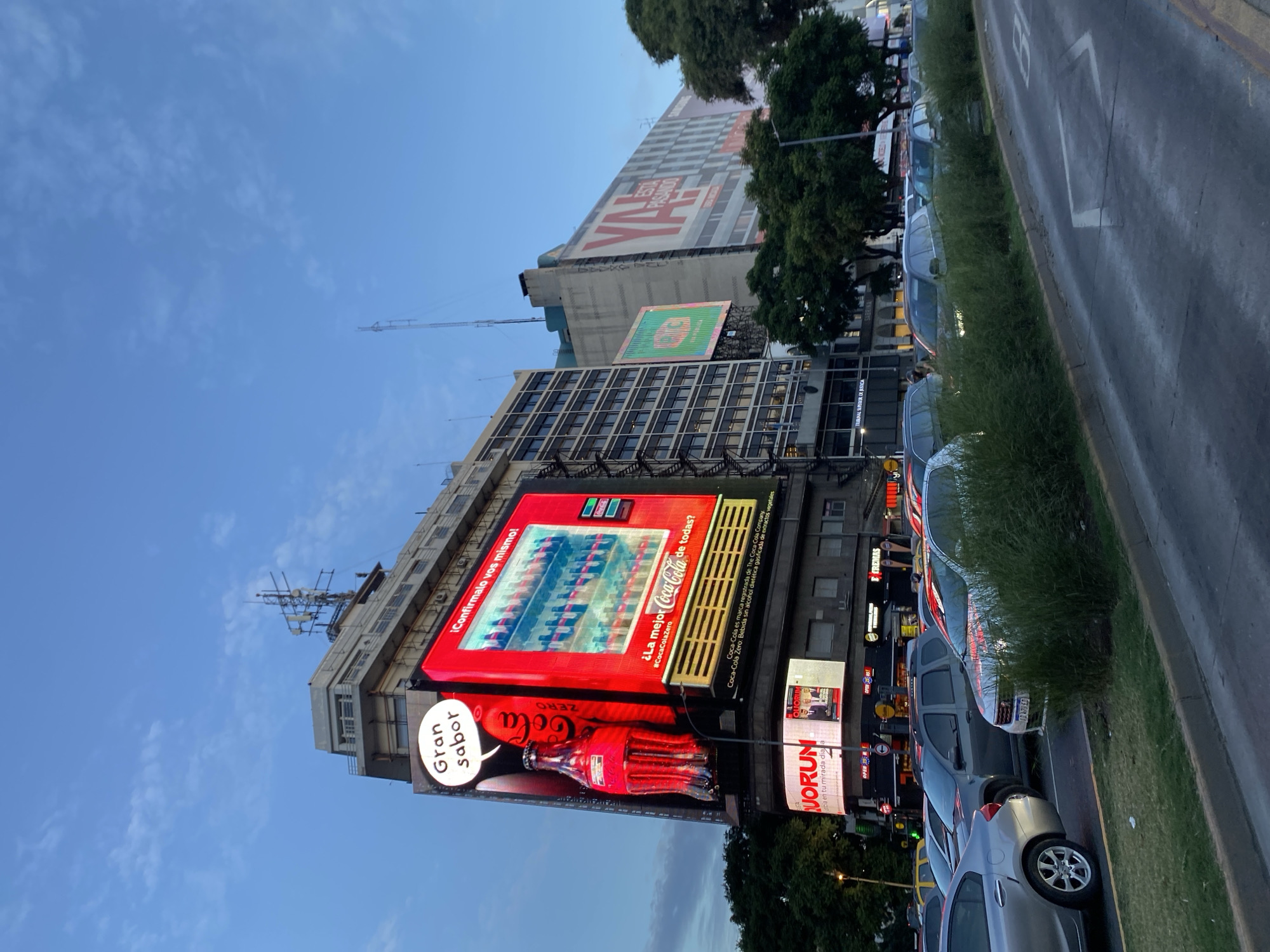 The image shows a street scene in Buenos Aires, Argentina, dominated by a building with a large Coca-Cola advertisement displayed on its facade. The ad features the phrase "Gran Sabor" (Great Flavor) and promotes Coca-Cola Zero. The advertisement is positioned above a smaller advertisement for "Quorum" which could be the name of a business or service. Cars are parked along the side of the street next to a grassy median. The sky is visible above the buildings, with clear patches intermixed with wispy clouds. Additional buildings are seen in the background.