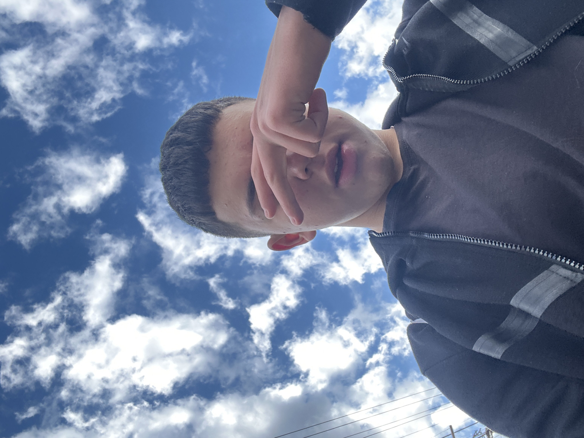 The image shows a person looking upwards towards the sky, partially obscuring their face with their hand. The sky is blue with scattered white clouds. The person is wearing a dark-colored jacket. The location appears to be outdoors, possibly in Zlatograd, Bulgaria, given the context of the prompt. The person is the main subject of the photo, with the sky and clouds providing the background.
