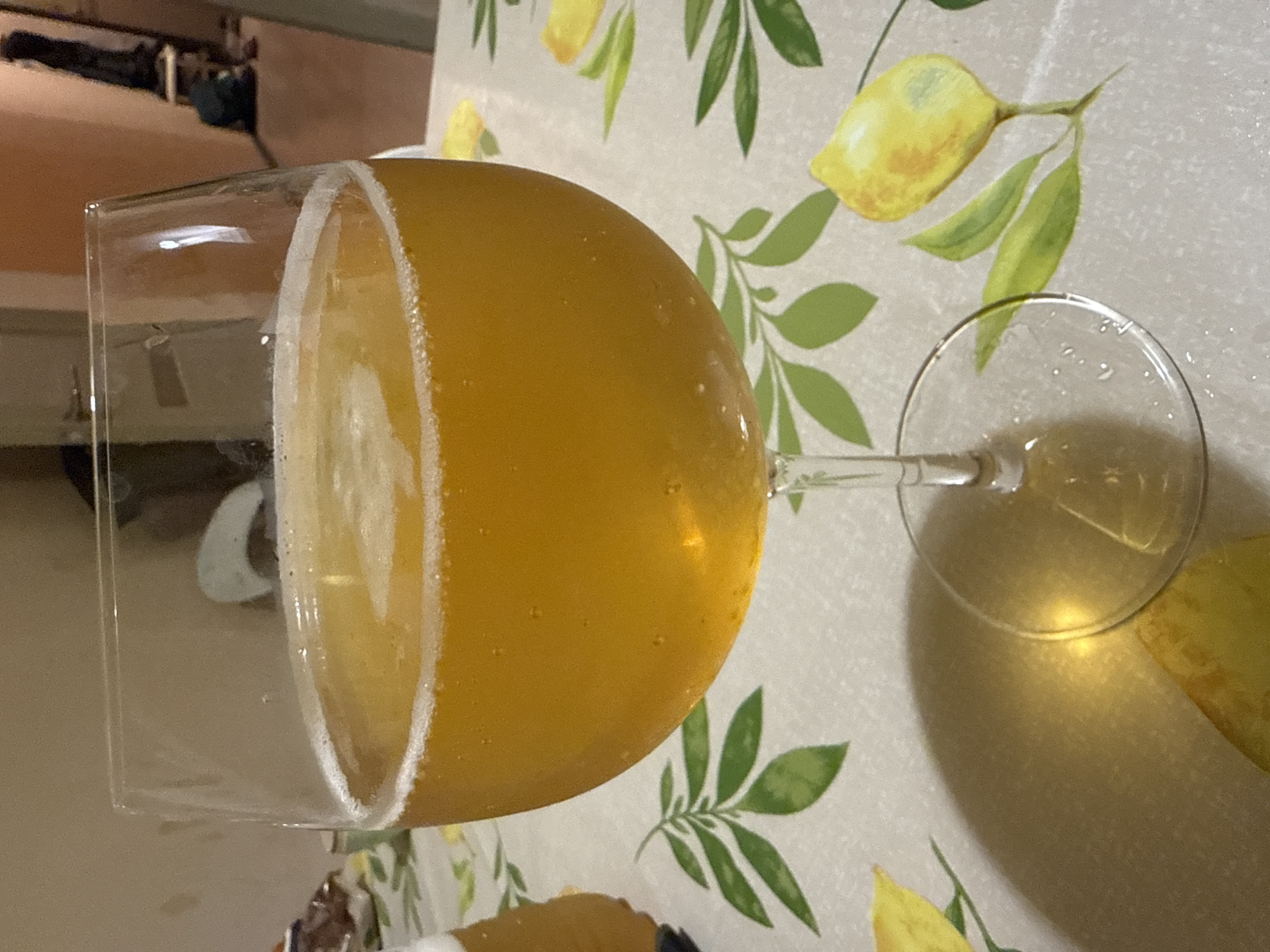 This close-up image, taken in Tobolsk, Russia, displays a large, wide-bowled glass, similar to a goblet, filled with a golden-yellow, translucent beverage. A thick, creamy white head of foam clings to the interior rim and sides of the glass, suggesting a carbonated drink like beer or cider. The glass is positioned horizontally, resting on a light-colored tablecloth.
The tablecloth features a repetitive pattern of yellow lemons with attached green leaves, adding a bright and decorative element to the scene. Reflections are visible on the glass, particularly on its stem and bowl, indicating an indoor setting with artificial or natural light. In the soft-focus background, indistinct shapes and darker tones can be perceived, possibly furniture or other objects within a room.
The image focuses primarily on the drink itself, emphasizing its color, clarity, and the texture of the foam. No specific human activity is explicitly shown, beyond the presence of the beverage. There is no visible text. The overall impression is one of a prepared drink in a casual indoor setting.