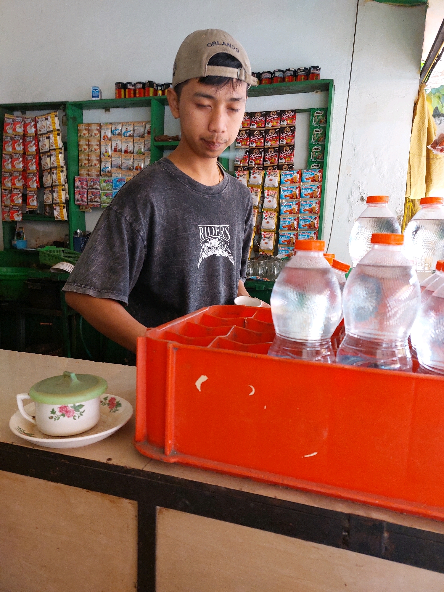 In Sumuragung, Indonesia, a young man wearing a light brown baseball cap with "ORLANDO" visible on the front and a dark grey, acid-washed t-shirt with a "RIDERS" graphic on the left chest stands behind a counter. He appears to be working in a small shop or convenience store.
The background reveals shelves stocked with various products. On the left, rows of small, colorful packets, likely snacks or instant food, are displayed. To the right of these, larger, multi-colored packages are stacked, featuring images of people and text. Above these, a shelf holds several small jars with colorful lids, possibly containing spices or condiments, and a blue box, possibly a cigarette pack.
In the foreground, on the counter, a small white teacup with a green lid rests on a matching saucer decorated with a floral pattern of pink roses. To the right of the teacup, a bright orange plastic crate holds several plastic bottles of clear liquid, presumably water, with orange caps. The counter itself is made of a light-colored material with a dark border. The overall environment suggests a humble, local establishment.