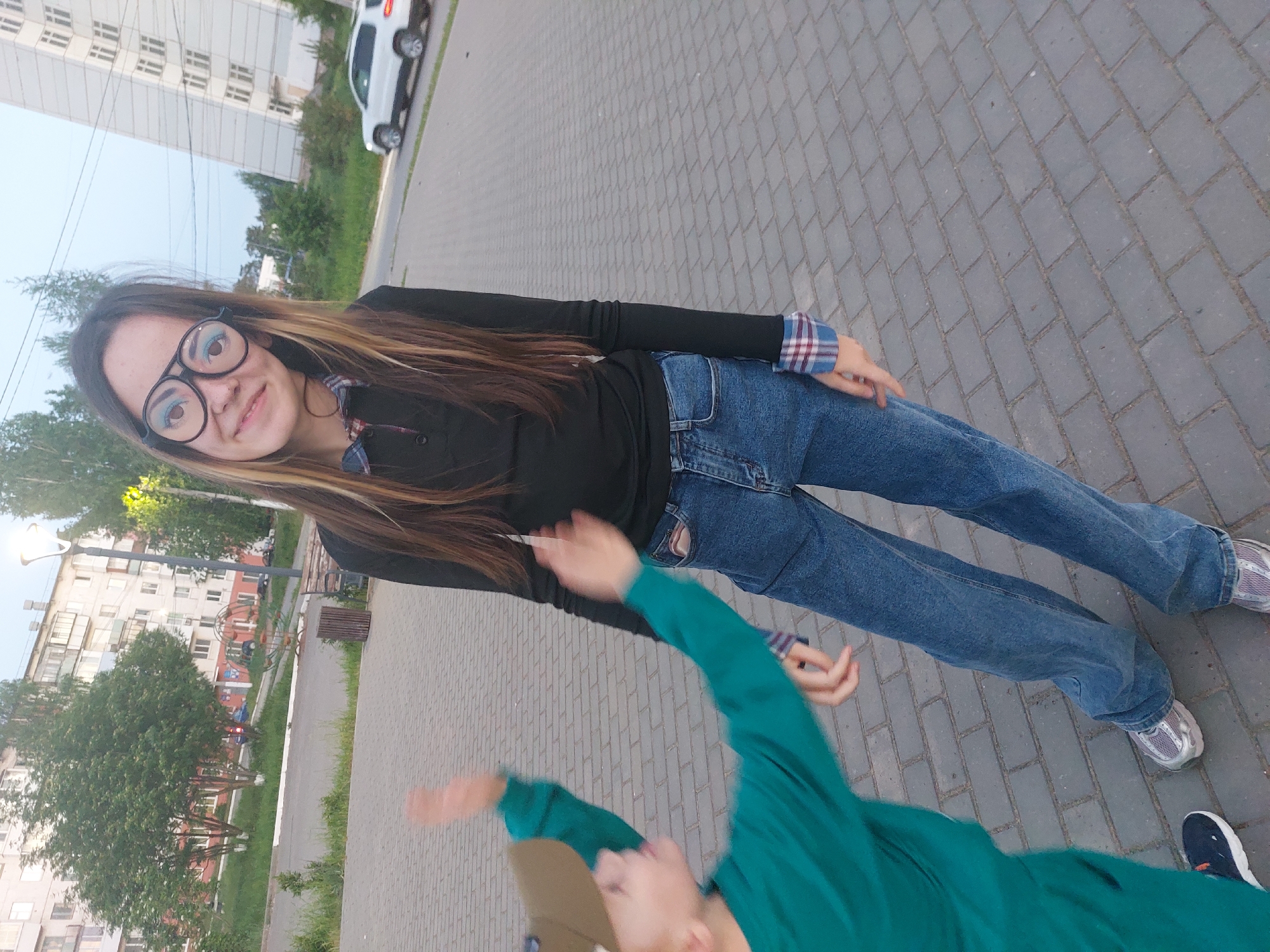 A young woman with long brown hair and large black glasses is standing on a gray brick sidewalk. She is wearing a black long-sleeved shirt with plaid cuffs and blue jeans. Her feet are shod in gray sneakers. In the foreground, a person is wearing a green long-sleeved shirt and is reaching up towards the woman. The background includes a tall building, trees, and a car. The location is Chaikovsky, Russia.