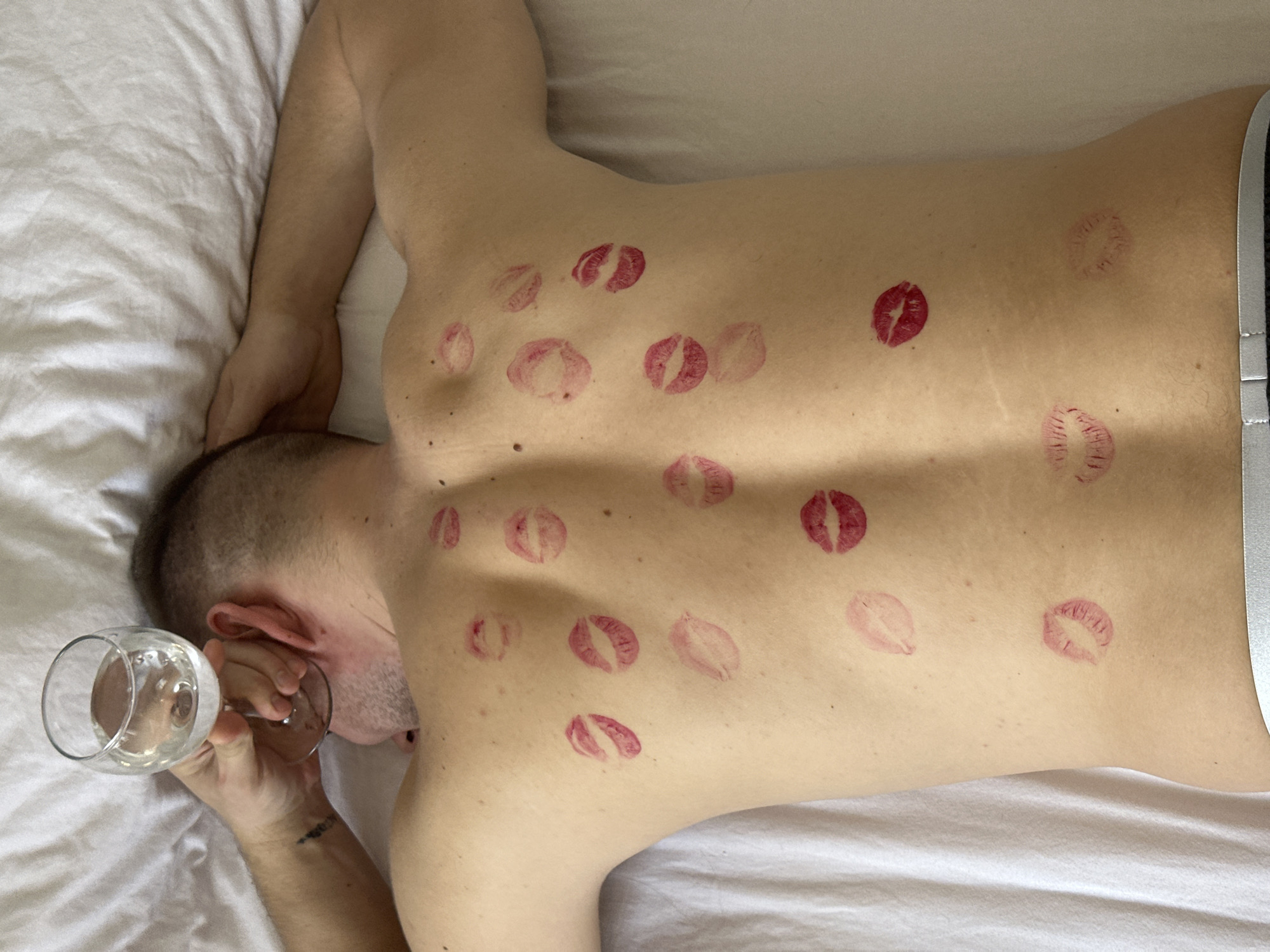 The image displays an individual, identified as male, lying prone on a bed covered with white sheets in Chaikovsky, Russia. The subject's bare back is predominantly featured, adorned with approximately 16-18 distinct red lipstick kiss marks scattered across the shoulders, back, and lower lumbar region. The head is turned to the left, and the right arm is bent with the hand positioned behind the head, grasping a clear stemmed glass containing a transparent liquid. A dark tattoo is partially visible on the inner right forearm. The left arm is extended above the head, out of frame. A grey waistband, consistent with underwear or shorts, is visible at the lower edge of the frame. The overall scene suggests an indoor, private setting.