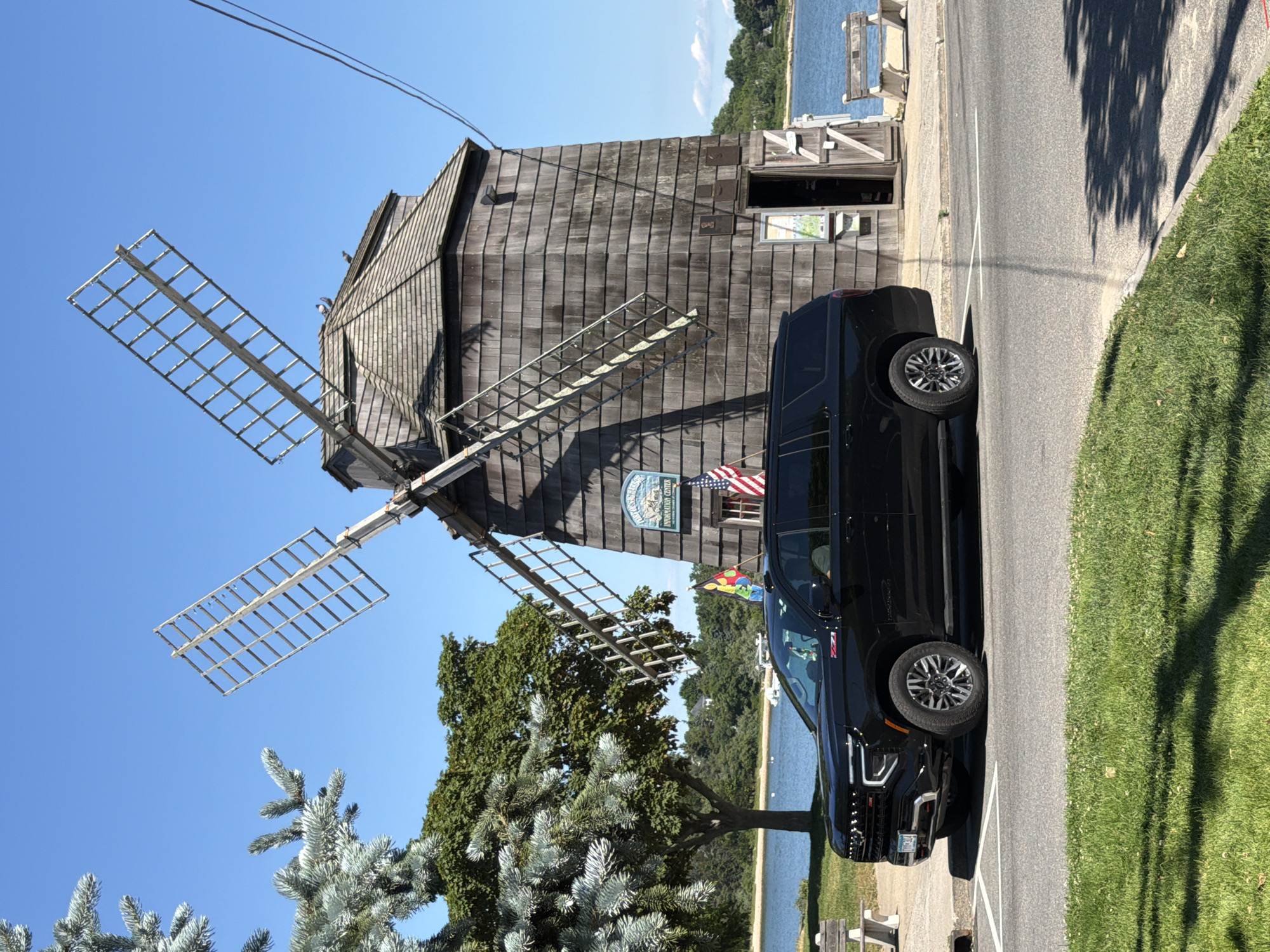 A black Chevrolet Suburban is parked on a paved road in front of a weathered wooden windmill. The windmill has two large, open wooden blades and a shingled roof. A sign on the windmill reads "Village of Sag Harbor Information Center." An American flag and a colorful flag are mounted on the windmill. A body of water, likely a bay or harbor, is visible behind the windmill, with trees and distant hills in the background. The sky is clear and blue. A patch of green grass is in the foreground, with shadows from trees cast upon it. There is a white bench to the left of the windmill. The license plate of the vehicle is partially visible and appears to be from New York.