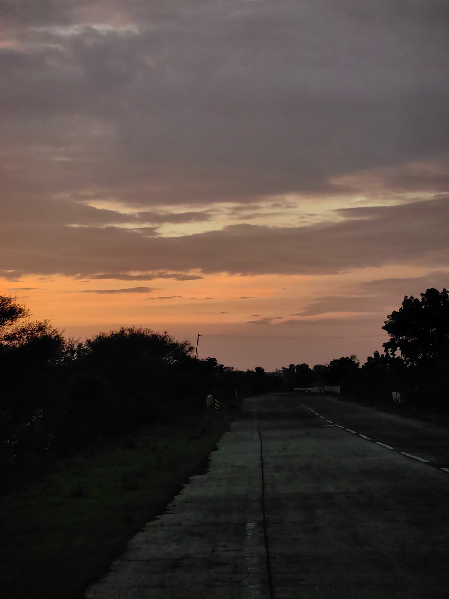 The image shows a rural road in Dattapur, India, at dusk. The sky is filled with dramatic clouds painted in shades of orange and purple, indicating the setting sun. The road, paved with asphalt and a concrete median, stretches into the distance, bordered by dark silhouettes of trees and bushes on either side. A few white markers or small walls are visible along the side of the road. The lighting is dim, suggesting either early morning or late evening. There are no people or vehicles visible in the scene.