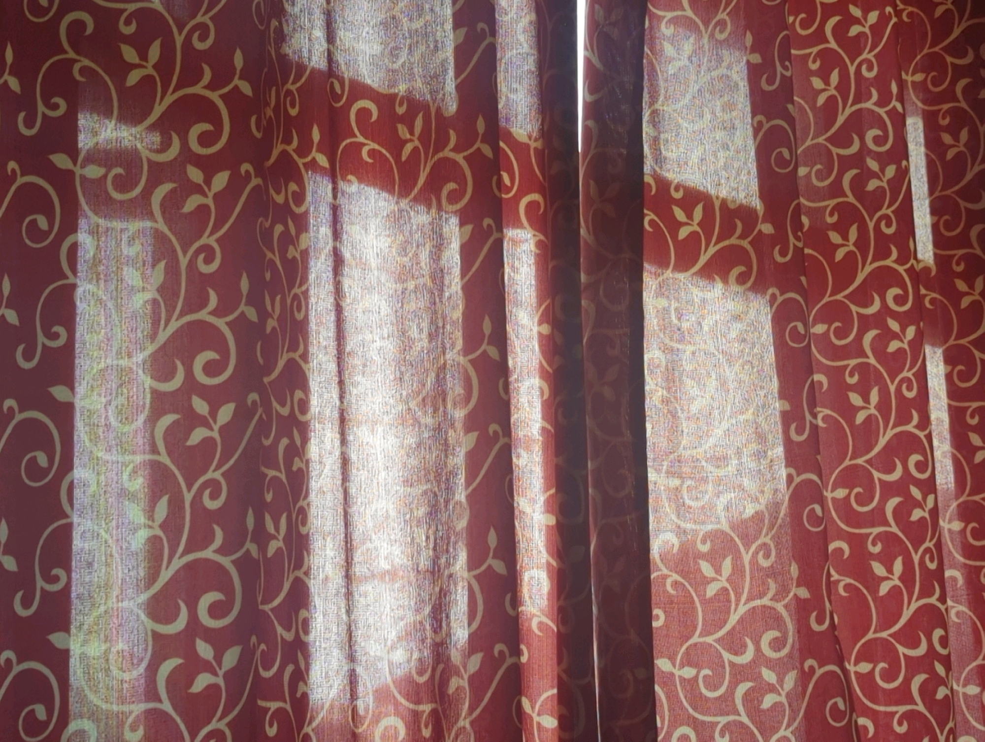 The image shows a close-up view of sheer red curtains with a light yellow vine pattern. The curtains are drawn, creating vertical folds. Sunlight is streaming through the fabric, casting bright, elongated rectangular shadows that suggest the presence of window blinds or a similar light-filtering structure behind the curtains. The texture of the fabric appears somewhat coarse and semi-transparent, allowing the light to diffuse through. The scene is indoors, likely a room, and given the context, it is situated in Baroo Tsok, India. There are no people or explicit objects other than the curtains and the light. The focus is on the interplay of light and shadow on the patterned fabric.